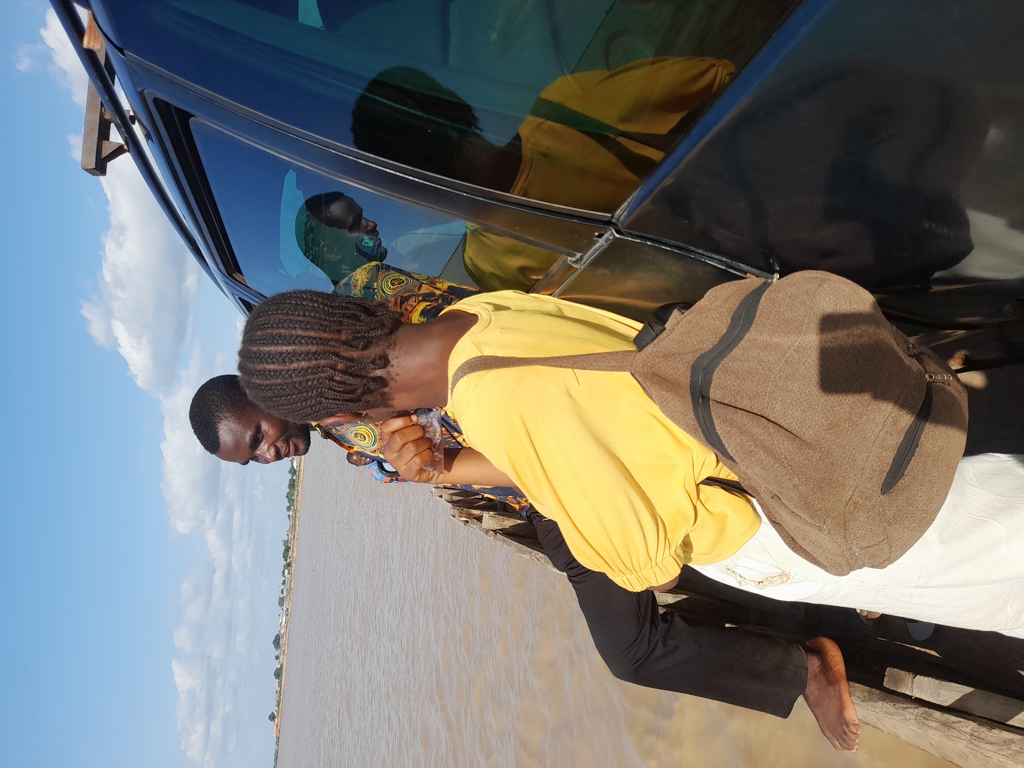 Two individuals are on a wooden boat navigating a wide, calm river. The person in the foreground, facing away from the camera, wears a bright yellow long-sleeved shirt and white pants. They have braided hair and are carrying a brown canvas backpack. To their left, a man with dark skin and short hair, wearing a patterned shirt, is seated. He is looking towards the foreground individual, with one hand resting on his leg and the other appearing to hold something. In the background, a reflection in the tinted window of a dark vehicle shows the man and another individual. A third man, wearing a blue and orange patterned shirt, is visible in the distance on the boat, looking towards the left. The environment is bright and sunny with a few scattered clouds in the blue sky. The riverbanks in the distance appear to have sparse vegetation and small settlements. The scene suggests a moment of travel or waiting on the river, likely in a rural or semi-urban setting. The location is Ibi, Nigeria.