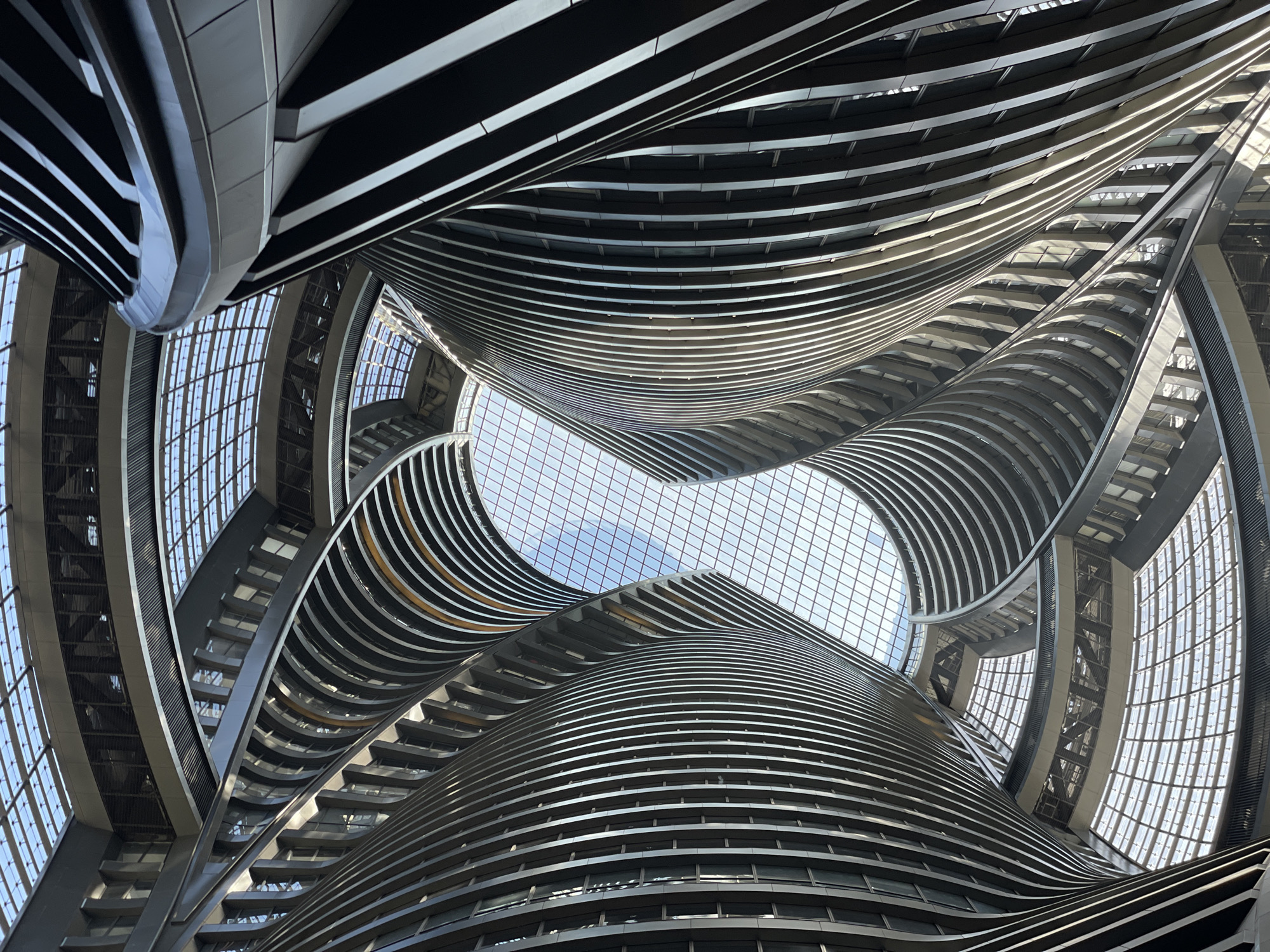 The image shows an upward-facing view of an interior architectural space. The building is characterized by a striking design featuring layered, curved balconies or decks that appear to spiral upwards towards a glazed opening, which reveals a view of the sky with light clouds. The structure exhibits a modern, steel and glass construction. The dominant colors are shades of gray and white, with the light from the sky adding brightness. The photograph appears to be taken from the inside of a building located in Fengtai District, China. The design is abstract and visually complex.