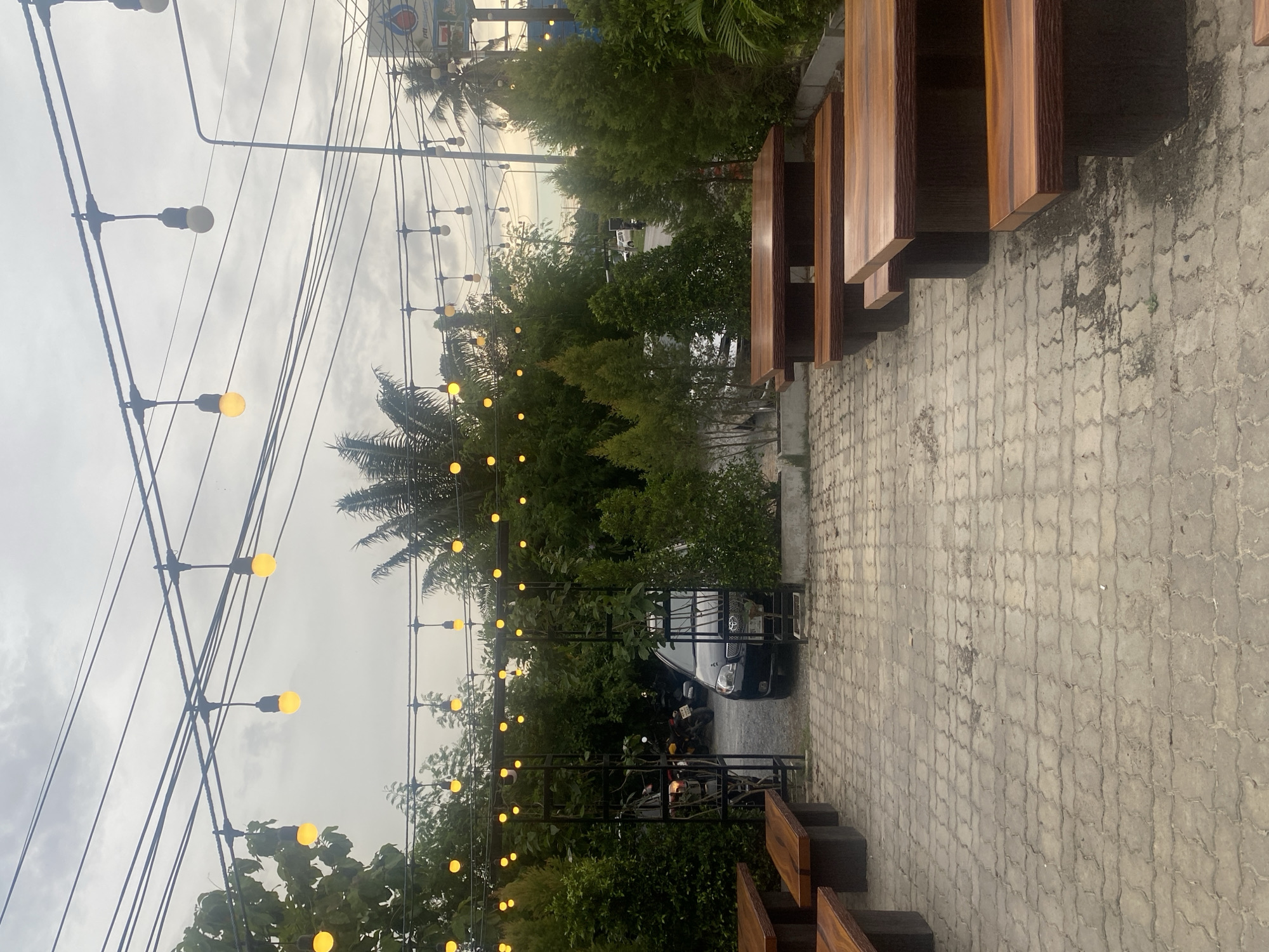 The image shows an outdoor dining area in Choeng Thale, Thailand. There are wooden tables and benches placed on a gray brick-paved area. Above, string lights with yellow bulbs are suspended between trees and metal posts. In the background, a black car and a few motorcycles are parked. The sky is overcast and several utility wires are visible. Green trees and foliage are present throughout the scene.