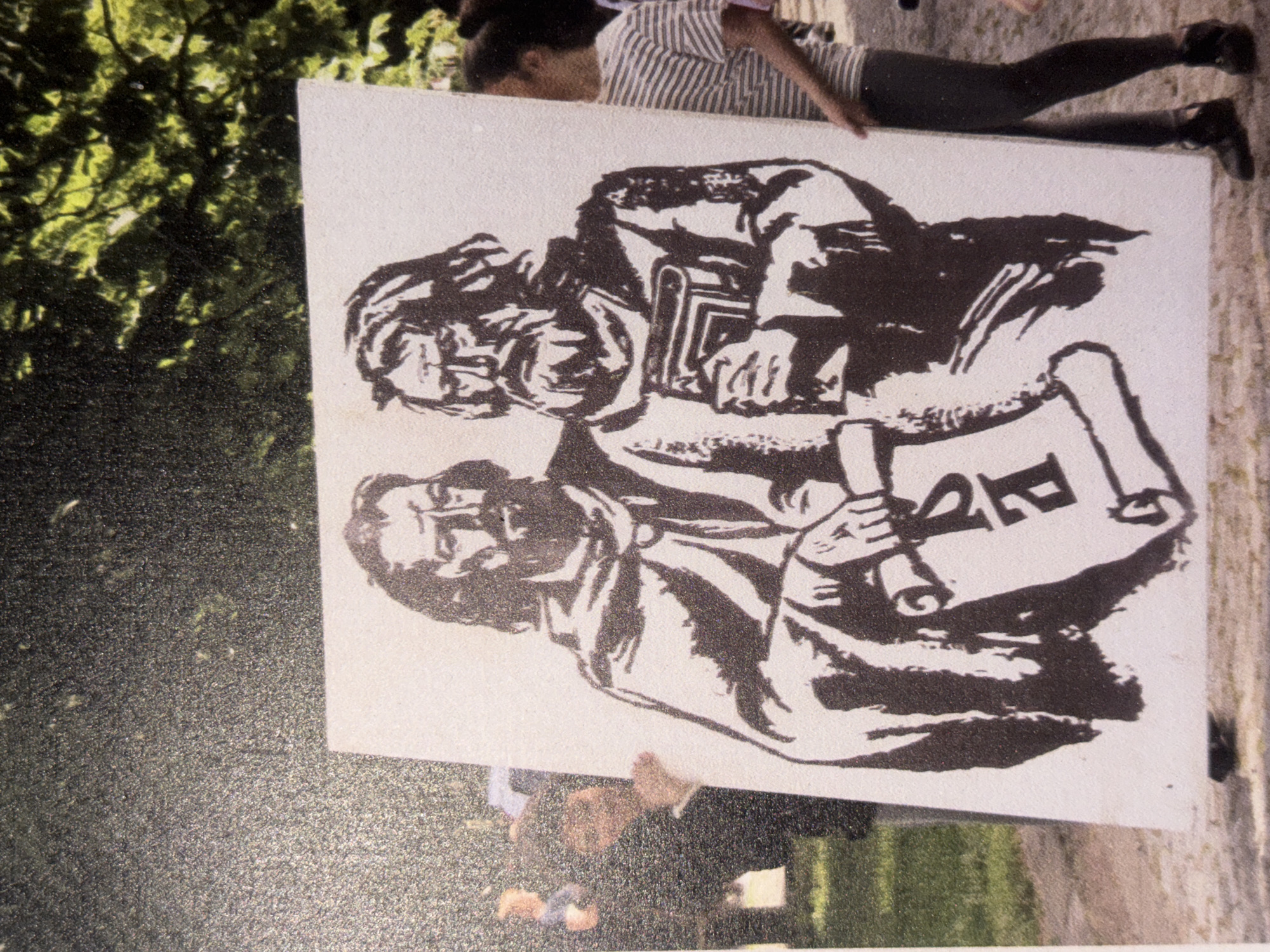 The image depicts an outdoor scene in daylight, focused on two individuals holding a large white sign vertically. The sign features a stylized black and white illustration of two bearded men, positioned one above the other, in a graphic, almost woodcut-like style. The upper man is shown holding a book, while the lower man holds an unfurled scroll. On this scroll, a prominent, stylized letter or symbol, resembling a capital "P" or possibly "PR," is visible.
One person holding the sign, positioned to the viewer's right side of the sign, is partially visible, wearing a striped shirt, dark trousers, and dark shoes. The other person, holding the left side of the sign, is largely obscured, with only parts of a dark jacket and hands visible. The background primarily consists of lush green foliage, suggesting a park or garden setting. Another person is partially visible in the lower right background, behind the sign. The image itself appears slightly grainy.