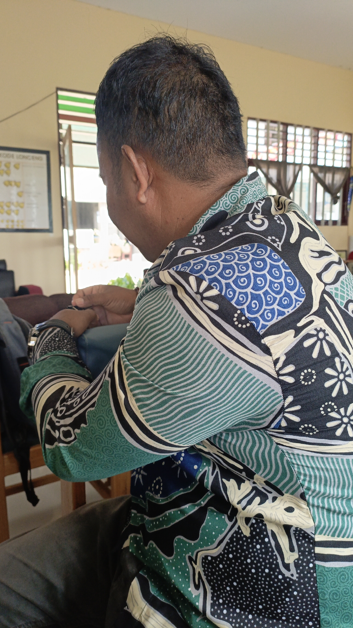 A medium shot captures a person from the back, viewed over the left shoulder, in what appears to be an indoor setting. The individual is wearing a long-sleeved batik shirt with a colorful pattern featuring green waves, blue swirls, and black and white floral designs. Their left arm is bent, with their hand resting on a dark blue object, possibly a chair or cushion. Their right arm is also bent, and their hand is visible near their chest. The person has short, dark hair and an ear is visible.
In the background, a pale yellow wall is visible. To the left, a framed chart with yellow markings is hanging on the wall, and a window with green and white horizontal stripes is present. To the right, a window with a grid pattern is visible, with dark curtains drawn. The lighting suggests an indoor environment with natural light coming from the windows. The overall impression is that of a casual, everyday scene. The image is static and lacks any clear indication of movement or dynamic interaction. The focus is primarily on the person and their attire.