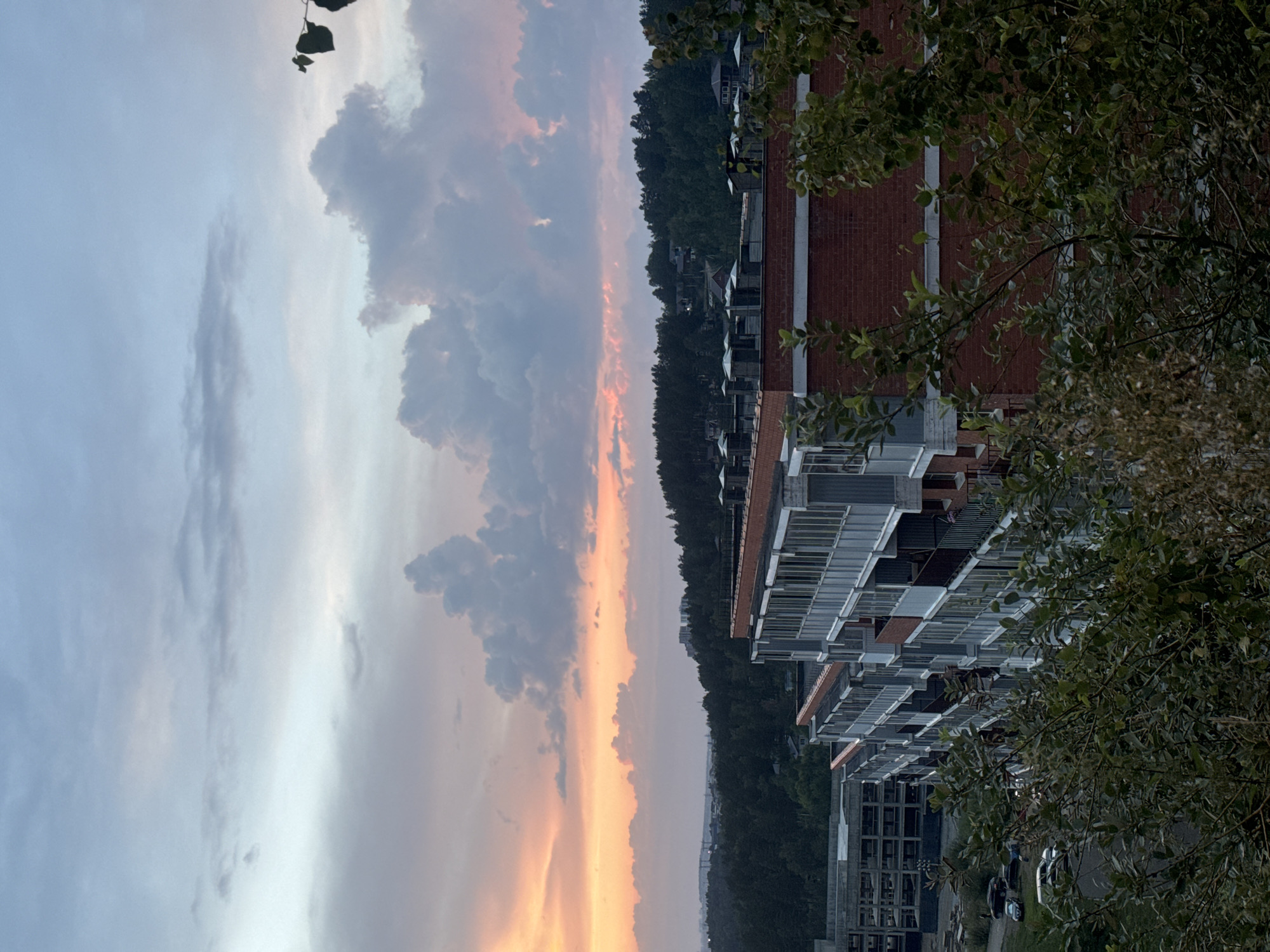The image shows a wide-angle view of a cityscape at dusk, with a focus on an apartment building. The sky is filled with clouds, some tinged with orange and pink from the setting sun, and others a soft grey. In the foreground, the top of a red brick apartment building with white-trimmed balconies is visible. Behind it, another, larger apartment building with a concrete exterior is under construction. A dense forest of green trees stretches across the middle ground, with some houses scattered within. The background shows a distant hazy cityscape, suggesting the location is Markova, Russia. The overall lighting suggests it is late evening. No people are visible in the frame.
