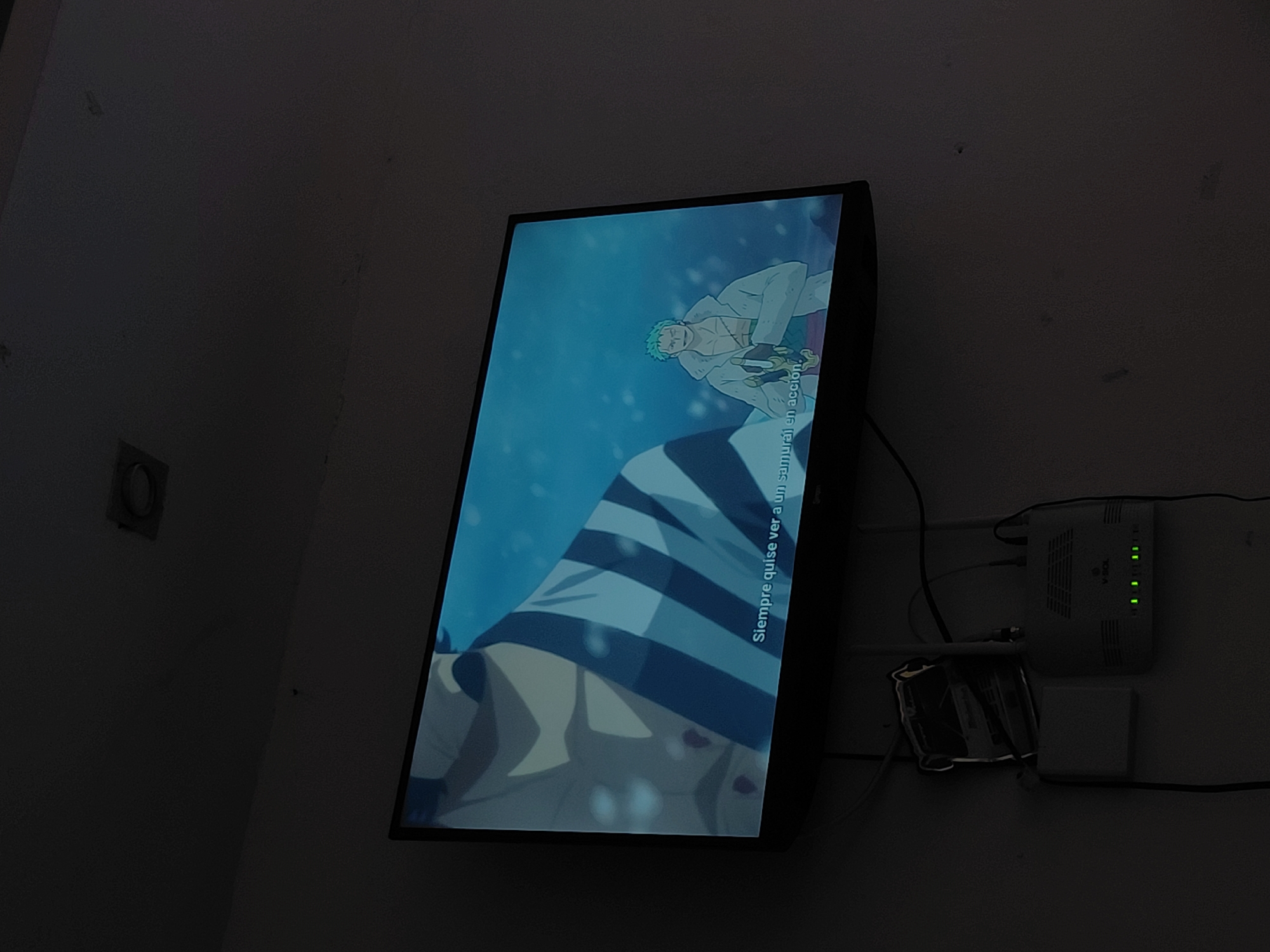 The image shows a television screen displaying an anime scene, likely from the show "One Piece," featuring characters Roronoa Zoro and another samurai. The television is mounted on a dark wall. Below the television, a white Wi-Fi router with several green indicator lights is visible, along with some cables. The overall lighting in the room is dim. The image was taken in Santa Ana, Venezuela.