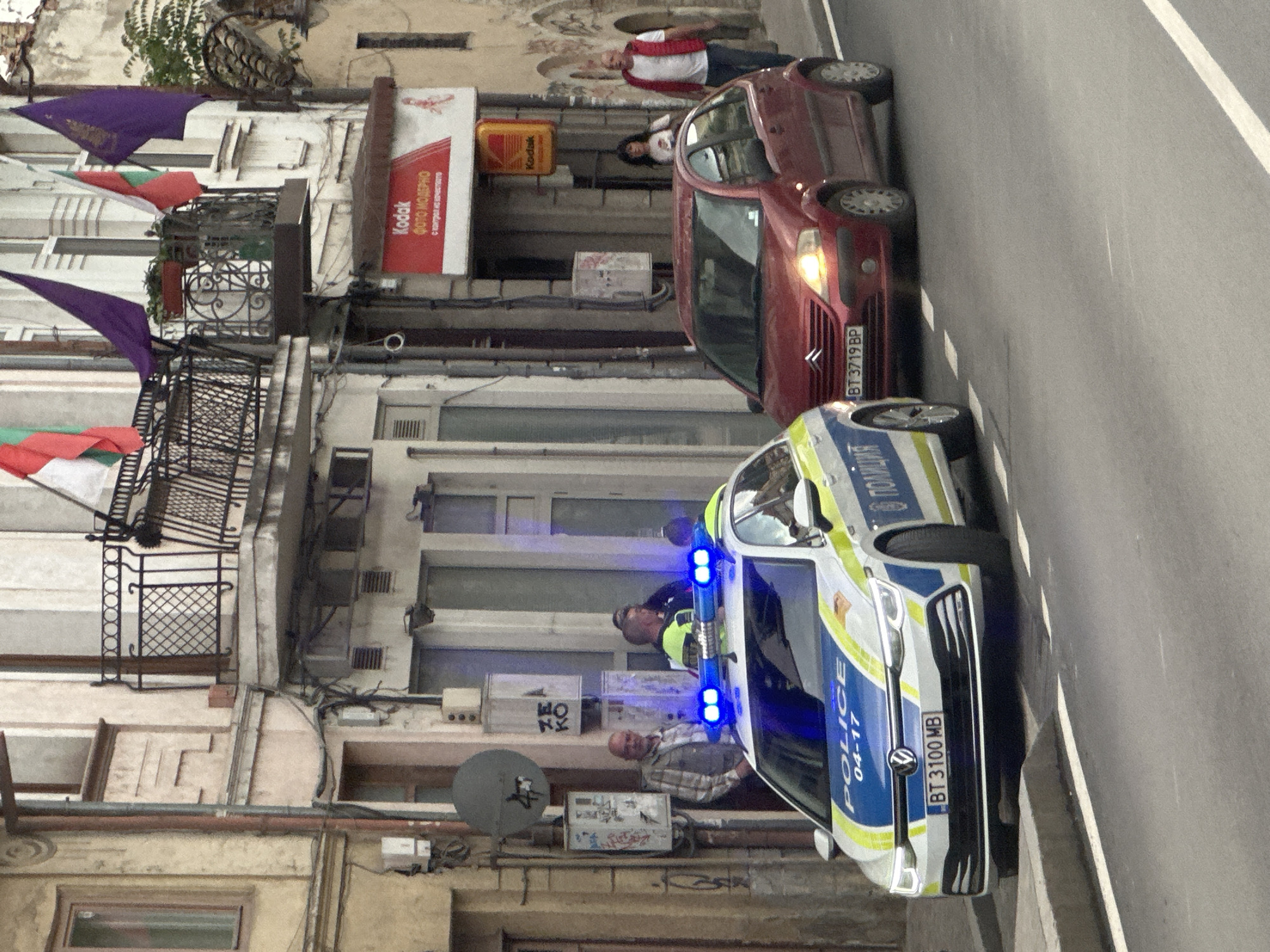 The image shows a street in Veliko Tarnovo, Bulgaria. A white and blue Bulgarian police car, with license plate BT 3100 MB and the markings "POLICE 04-17" and "В полиция," is parked on the right side of the street with its blue lights activated. In front of the police car is a red Citroen car, license plate BT 3719 BP, parked in the opposite direction. Several people are visible on the sidewalk and in the background. The buildings on the left side of the street are adorned with Bulgarian flags and purple pennants. A Kodak photo shop sign is visible on one of the buildings. A man in a red vest stands near the red car. Two additional men stand next to the police car. A satellite dish is mounted on the building.