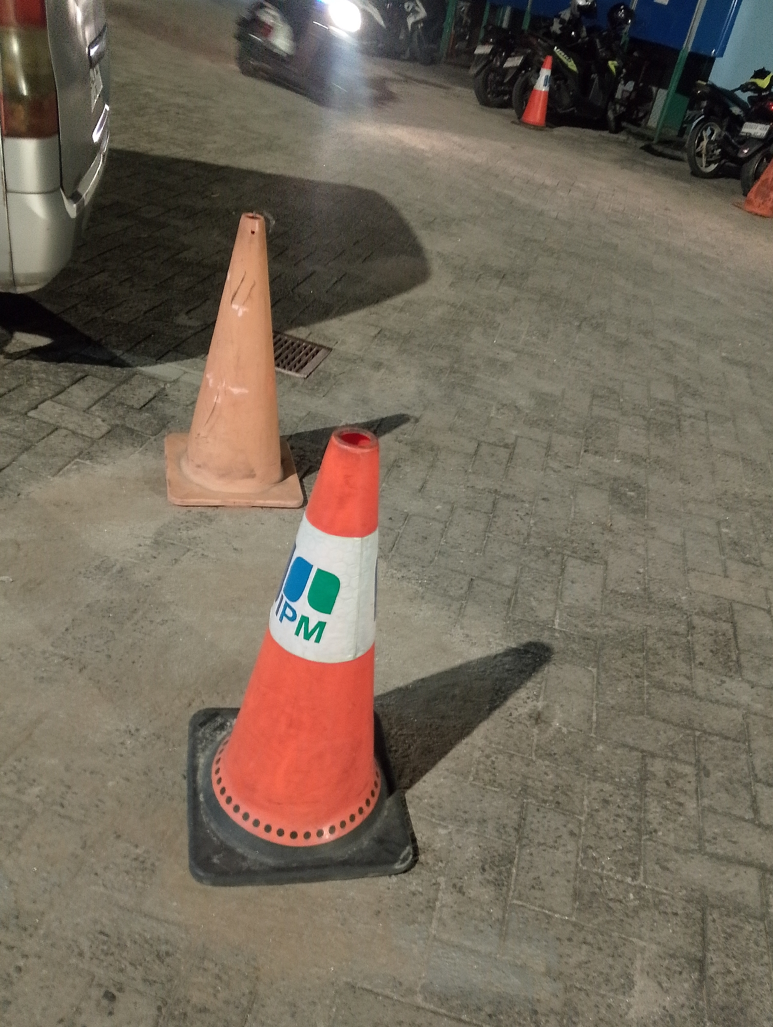 The image depicts an outdoor scene at night, likely in the City of Pematang Siantar, Indonesia, given the context. In the foreground, two orange traffic cones are prominently displayed on a paved surface. The cone on the right is closer to the viewer and features a white reflective band with blue and green logos that appear to read "IPM". The cone on the left is further back and appears to be a standard orange traffic cone.
Behind the cones, a portion of a silver vehicle is visible on the left side of the frame. In the background, several motorcycles are parked in a row. The lighting suggests artificial illumination, possibly from streetlights or building lights, casting shadows on the ground. The overall environment appears to be a parking area or a street. There are no people clearly visible in the image.