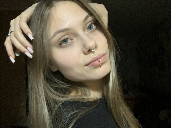 A young Caucasian female is depicted in a close-up, head-and-shoulders shot. She possesses long, straight, light blonde and brown ombre hair, styled with a side part, which extends over her shoulders. Her eyes are light blue, and she maintains direct eye contact with the viewer, exhibiting a slight smile. Notable facial features include a silver V-shaped septum piercing and two silver ball stud piercings positioned horizontally on her upper lip (philtrum). Her left hand is raised and rests on the top of her head, with a silver ring visible on her ring finger. Her fingernails are long, with a light pink base and white tips, consistent with a French manicure. She is wearing a black garment, likely a top or t-shirt. The background is uniformly dark and indistinct, suggesting an indoor setting with the subject well-lit from the front. The scene is located in the city of Foki, Russia.