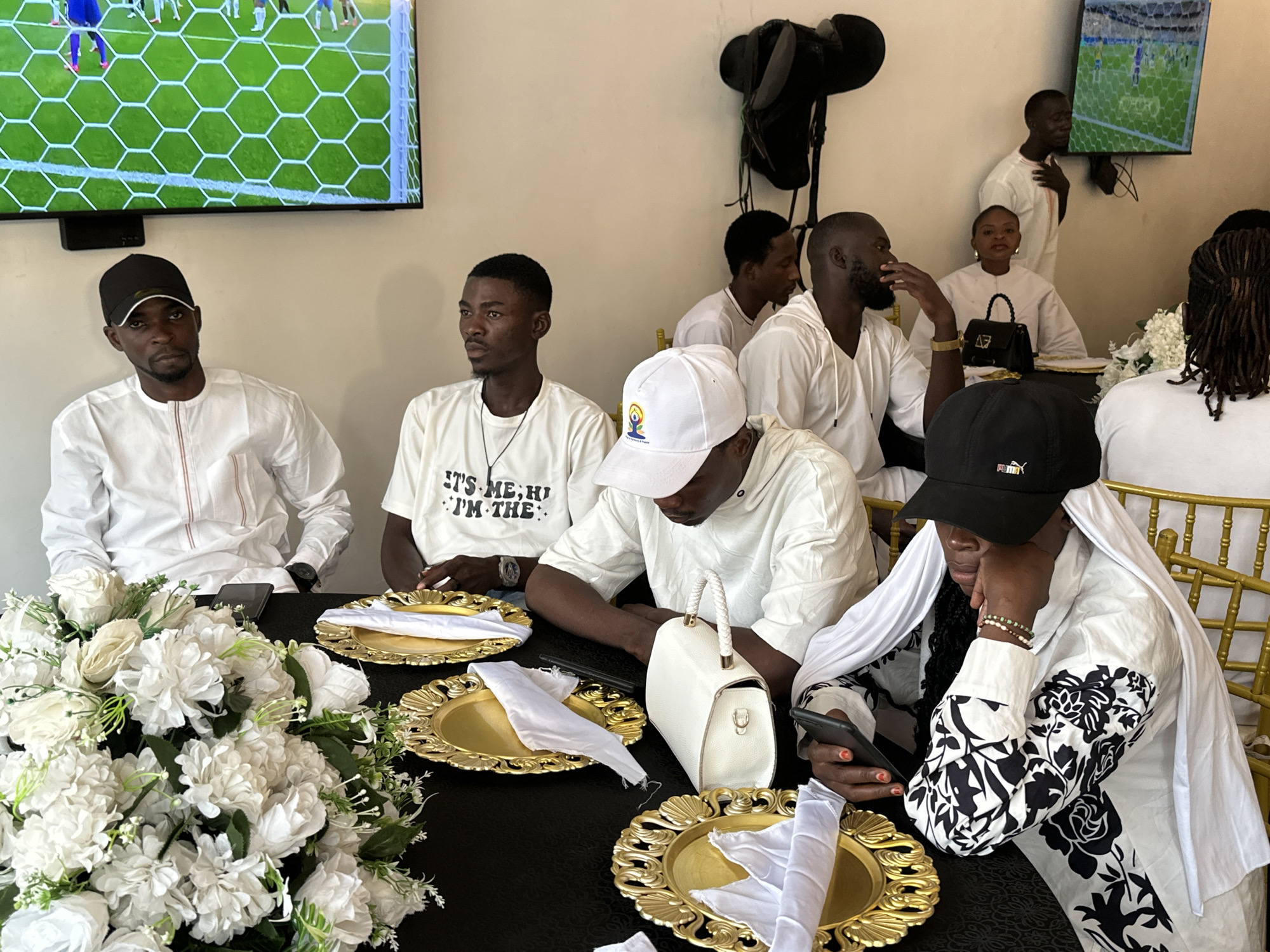 The image depicts a gathering of people seated at a table, seemingly for a celebratory event or meal. The scene is indoors, with a large television screen displaying a soccer match in the background. The table is adorned with white floral arrangements and ornate gold plates with white napkins. Several individuals are dressed in white attire, common for celebratory occasions in Nigeria.
The subjects include a group of men and at least one woman. Some men are wearing white shirts and caps, while others are in white hooded garments. The woman in the foreground is wearing a white garment with a black floral pattern and a white head covering, and is looking at her phone. A man standing in the background is also wearing white.
The atmosphere appears relaxed, with some individuals engaged in conversation or looking at their phones, while others are focused on the television. The lighting suggests it is daytime. The presence of the soccer match on screen might indicate a shared interest or a specific occasion related to a sports event.
Visible text includes "IT'S ME, HI! I'M THE:" printed on a t-shirt worn by one of the men. The logo on the cap of the person in the foreground, partially obscured, appears to be a "Puma" logo.