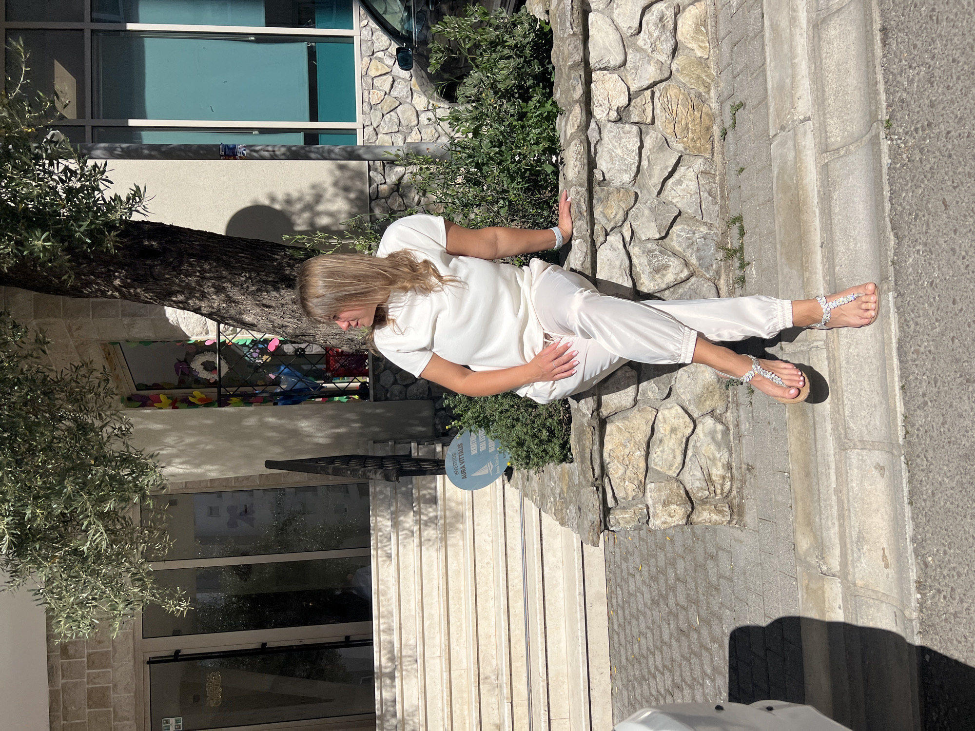 A woman wearing a white pajama set and sandals with sparkling embellishments is sitting on a ledge in front of a stone wall. She has blond hair and is wearing a wristband. The location appears to be in Budva, Montenegro. In the background, there's a building with windows and a tree. Steps lead up to the building. The area has stone elements in the architecture.