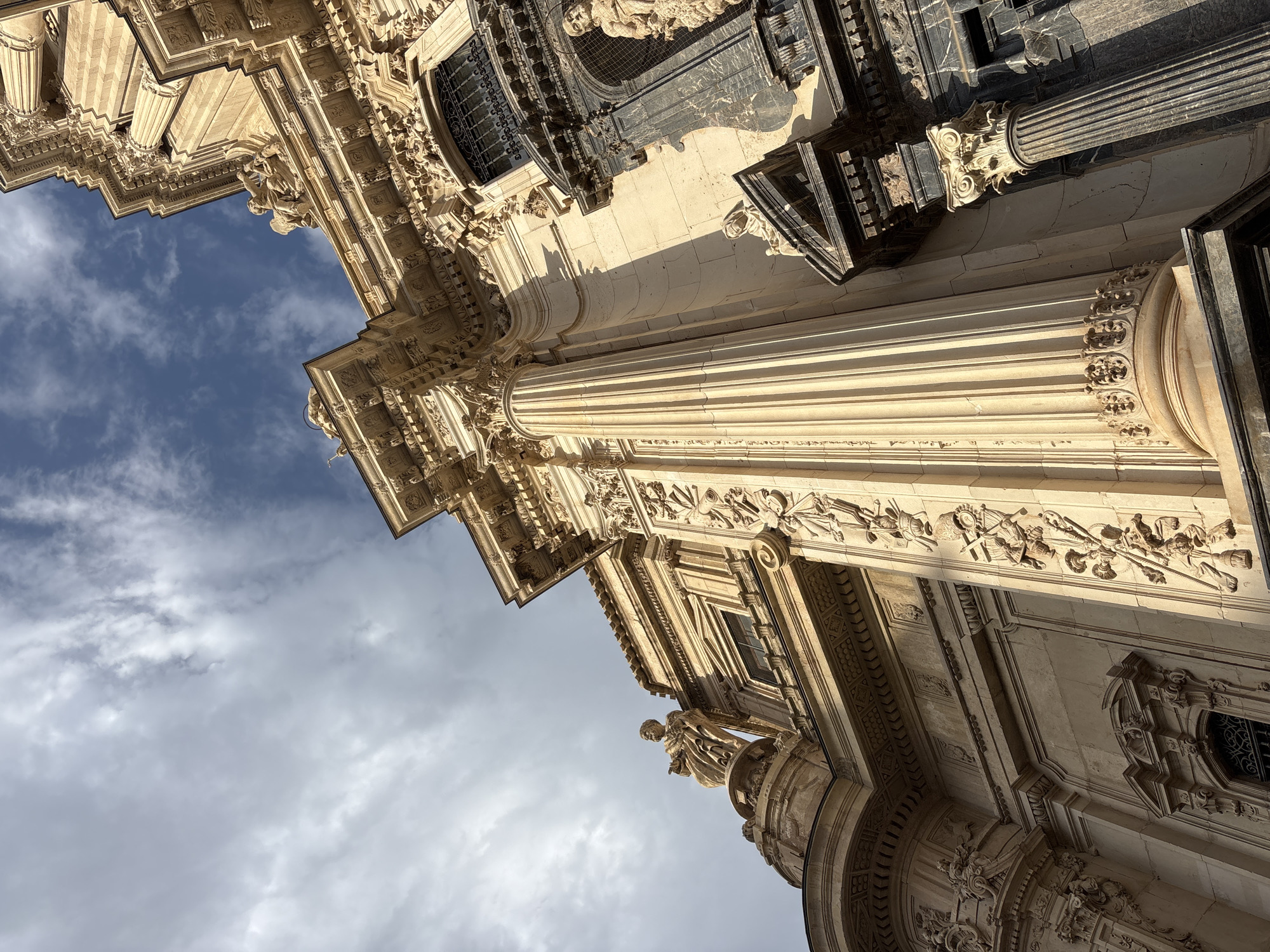 A low-angle, worm's-eye view captures the ornate Baroque facade of the Cathedral of Murcia, Spain, against a partially cloudy sky. The building's cream-colored stone is carved with intricate reliefs, including figures, architectural elements, and decorative motifs. A large, fluted Corinthian column dominates the foreground, casting a shadow across the textured stone wall. To the right, a darker, marble-clad section of the facade features another column and a niche with a statue. Sculptural details are visible throughout, including figures adorning the cornices and ledges. The sunlight highlights the three-dimensional textures of the carvings.