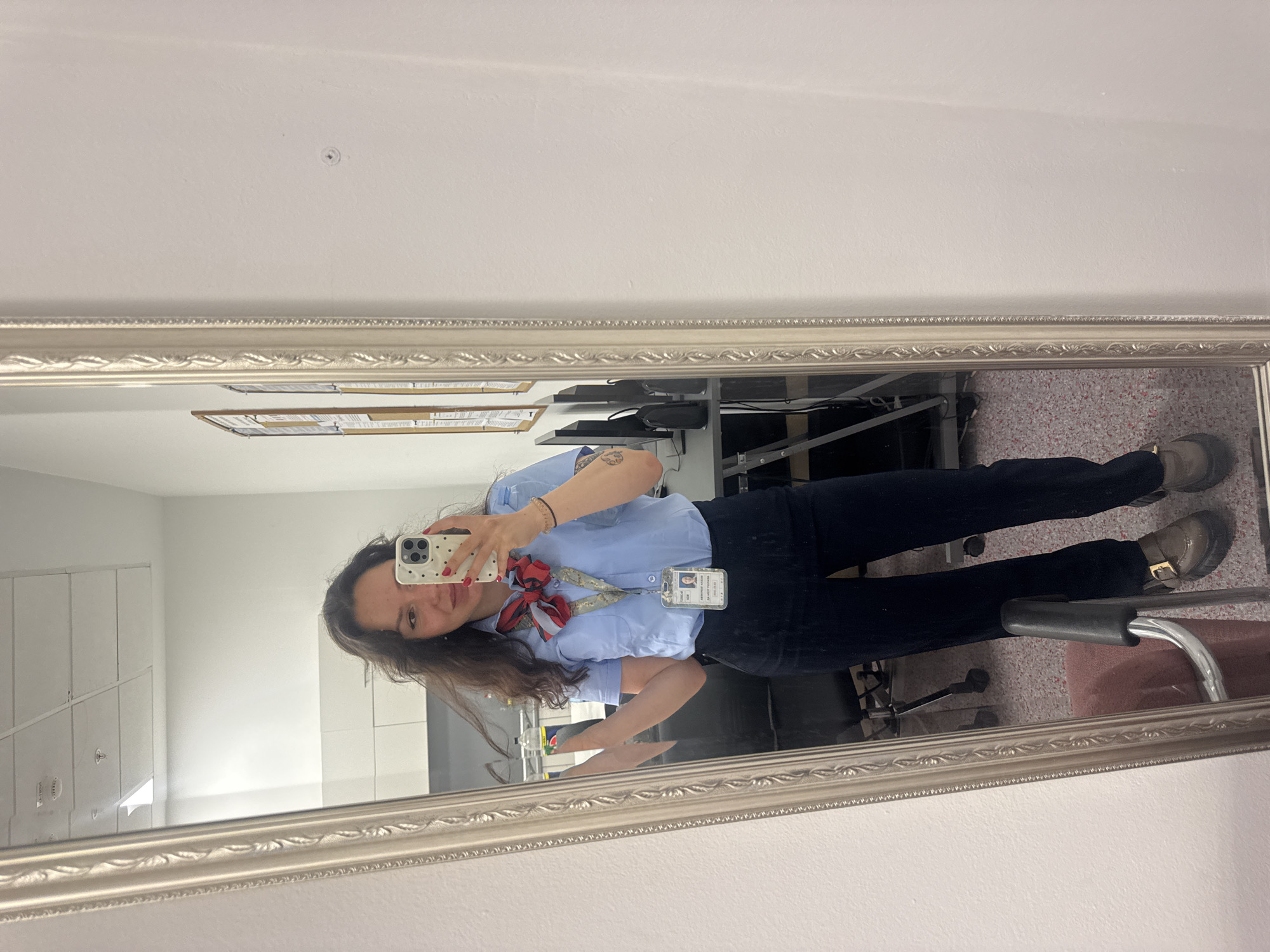 A young woman in a blue uniform shirt with a red and white neck scarf and dark pants is taking a selfie in a mirror. She is holding a white phone with a spotted case. The background, visible in the mirror, appears to be an office or workplace environment. The location is likely in Burgas, Bulgaria.