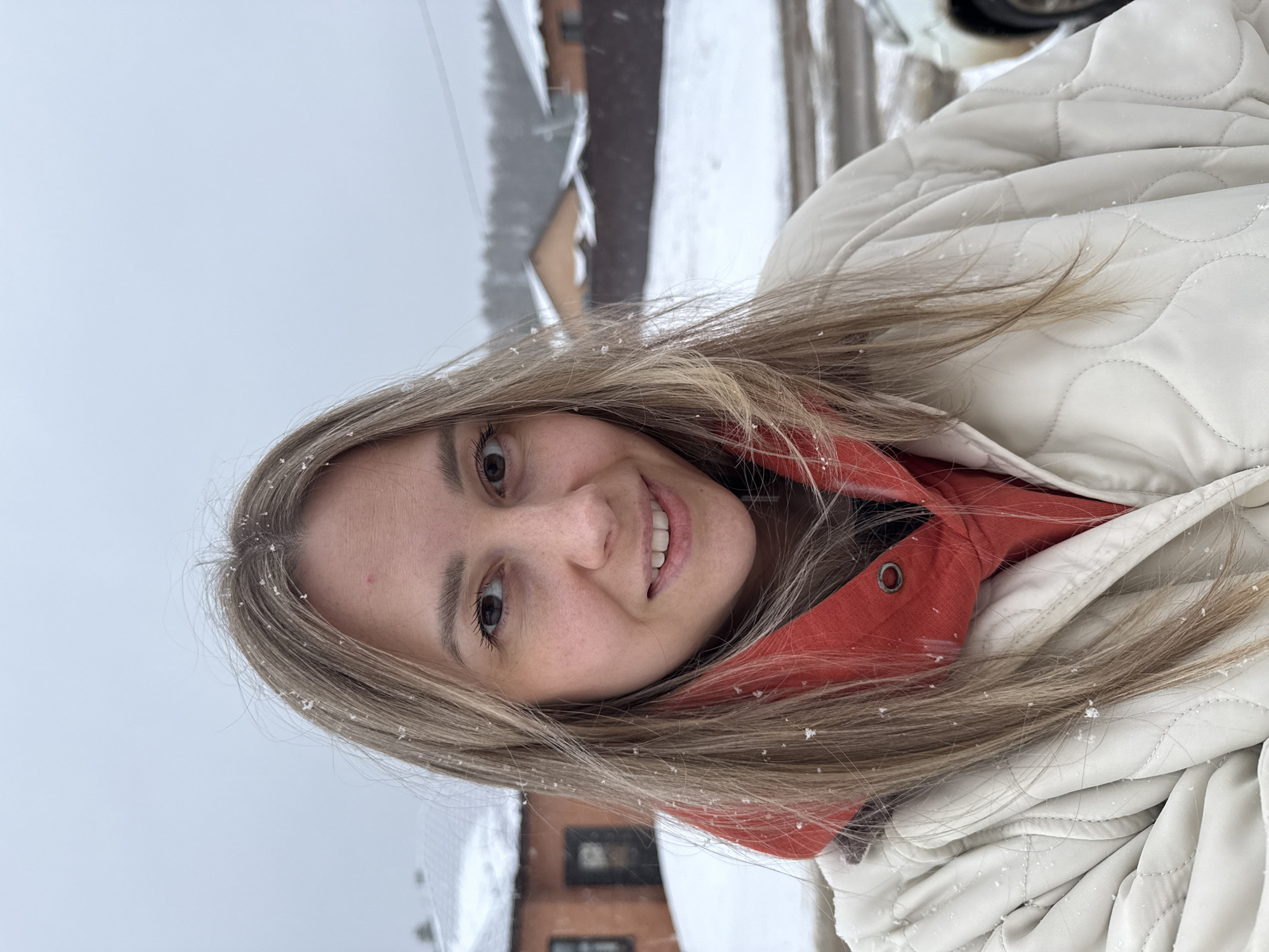 A young woman smiles at the camera while outside in a snowy environment. She has light brown hair with snowflakes resting on it, fair skin, and wears a red hooded sweatshirt under a cream-colored quilted jacket. In the blurred background, a building with snow-covered eaves is visible, suggesting a location in or near the city of Chaikovsky, Russia. Additional snowy terrain is also discernible in the background.