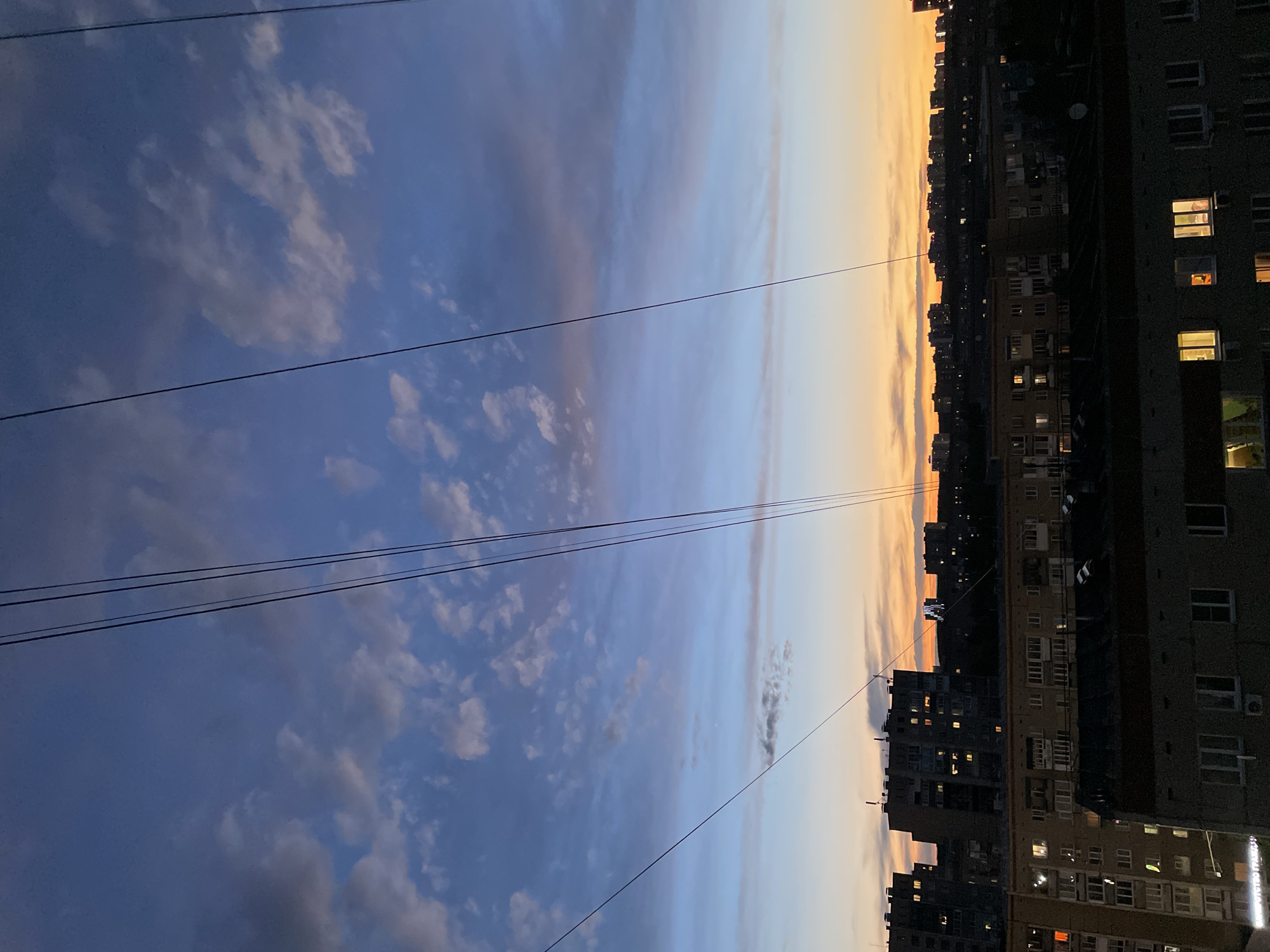The image shows a view of a city at dusk or dawn. In the foreground, there are power lines stretching across the sky. The sky is a gradient of colors, ranging from blue with scattered clouds on the left to orange on the right, indicating the presence of the setting or rising sun. On the right side of the image, there are several multistory apartment buildings. Some of the windows in the buildings are lit, suggesting people are inside. Based on the architectural style of the buildings, and particularly the presence of Cyrillic script on a sign on one of the buildings, this image is likely taken in Izhevsk, Russia. There are no people visible in the image. The focus of the image is on the contrast between the natural light in the sky and the artificial light from the buildings.