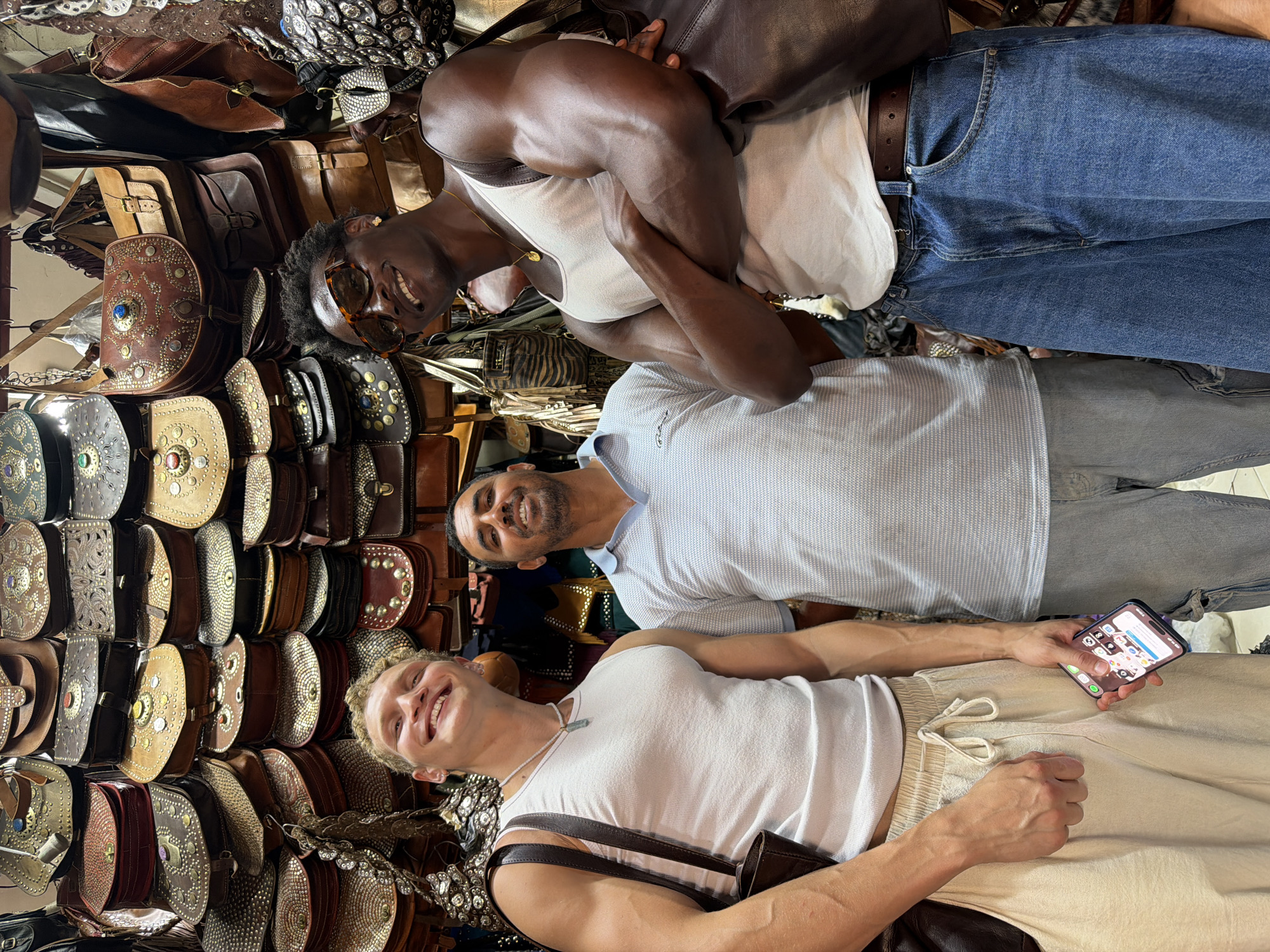 Three men are standing in front of a display of leather bags in a shop in Marrakesh, Morocco. The man on the left, who is white, is wearing a white tank top and off-white drawstring pants. He is holding a smartphone in his right hand. The man in the middle, who is of North African descent, is wearing a light blue and white striped polo shirt and light blue jeans. He is smiling and looking towards the camera. The man on the right, who is Black, is wearing a white tank top, dark sunglasses, and blue jeans. He is smiling and has his arms crossed. The background is filled with various styles of intricately designed leather bags.