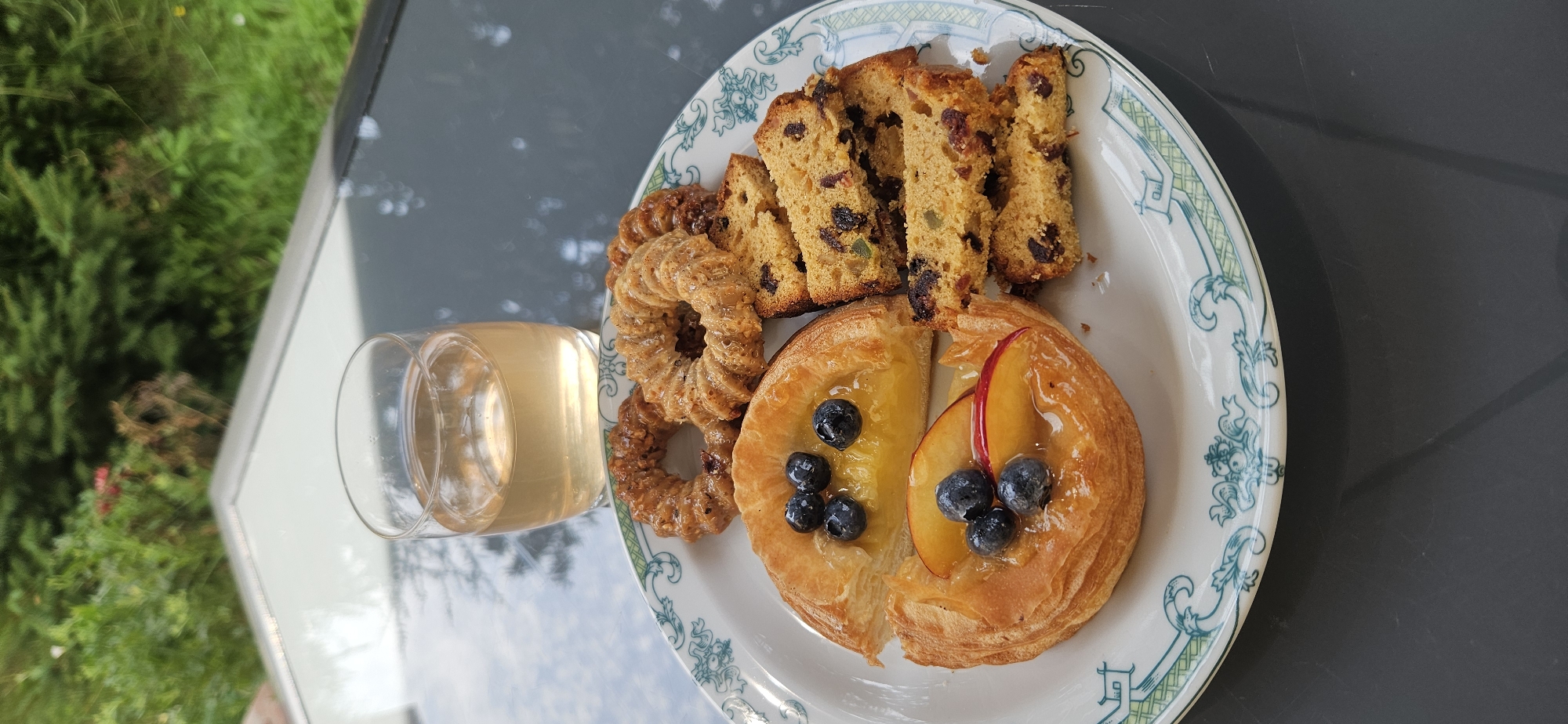 A plate of pastries and a glass of white wine are arranged on a dark, reflective surface, possibly a table, with a blurry green, leafy background suggesting an outdoor setting. On the plate, there are two ring-shaped pastries with a textured, glazed exterior, and several slices of fruitcake containing visible dried fruit and candied peel. Also on the plate are two danish pastries; one is a half-circle filled with a yellow, custard-like substance and topped with blueberries, and the other is a more irregularly shaped pastry filled with sliced peaches and blueberries, all glistening with glaze. The glass contains a light-colored liquid, consistent with white wine. The setting appears to be a patio or garden area, possibly at a cafe or restaurant in Hévíz, Hungary, given the presence of a scenic outdoor environment and a selection of baked goods and wine.