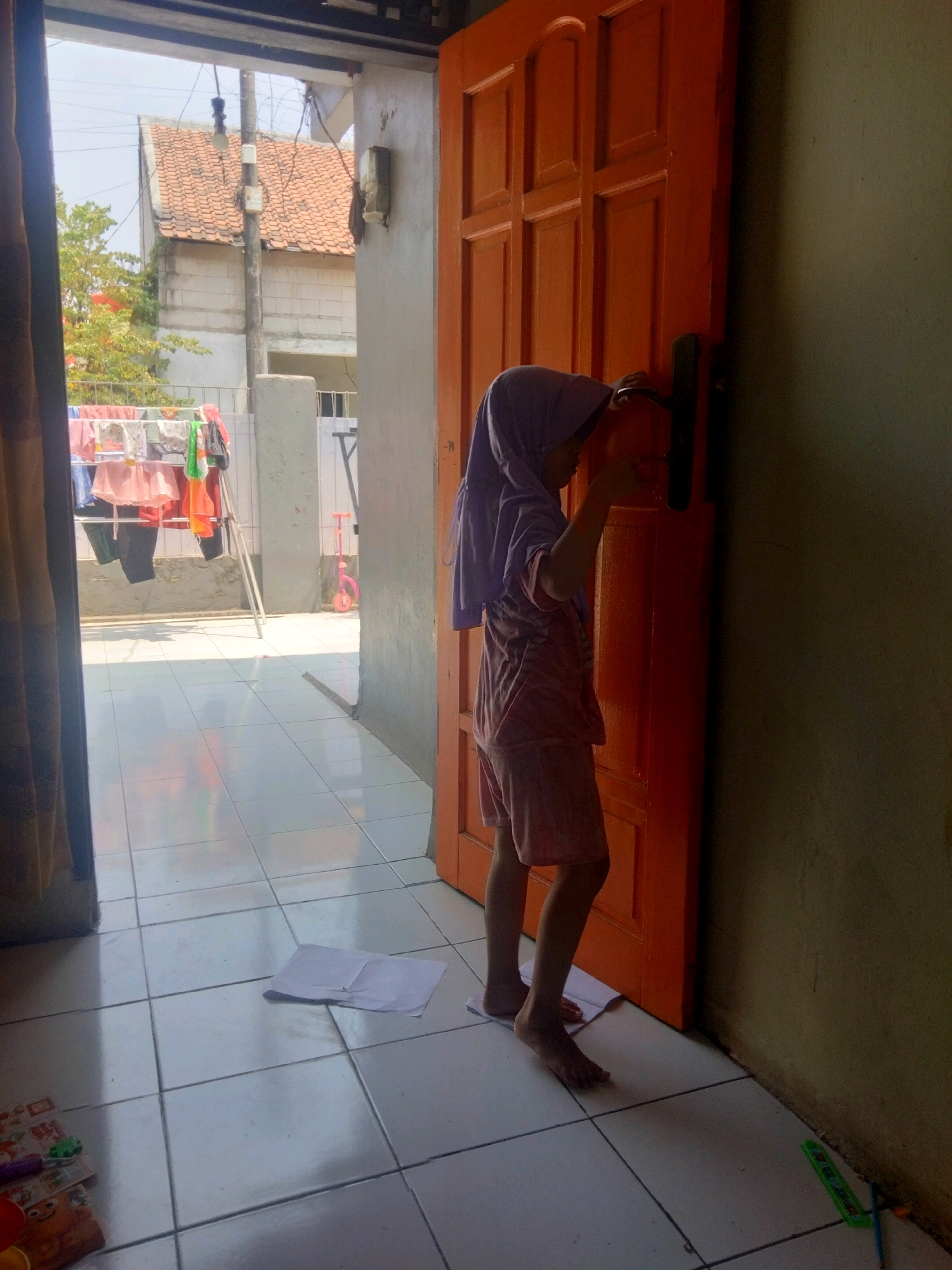 A young child wearing a purple hijab and a pink t-shirt and shorts stands on tiptoes in a doorway, reaching for the doorknob of a bright orange wooden door. The child's feet are bare, and one foot is placed on a white sheet of paper. The scene appears to be outdoors, likely in the yard of a house in Suka Sari, Indonesia, with a tiled floor reflecting sunlight. To the left, a clothesline is visible with laundry hanging, and a pink toy scooter is parked nearby. The background shows a building with a tiled roof and utility poles. The overall environment suggests a residential setting on a bright, sunny day.