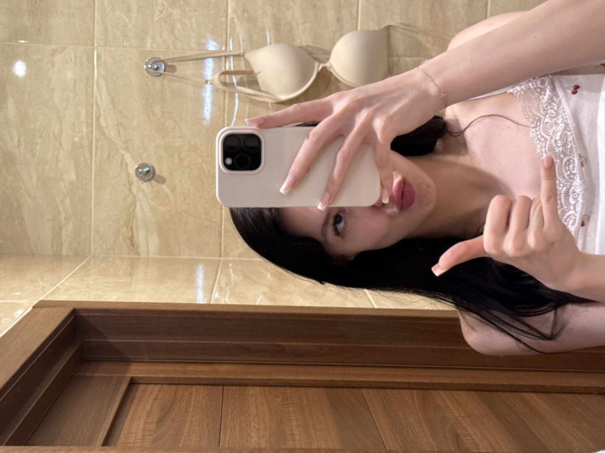 An undated, eye-level, medium shot shows a young woman with long, dark hair taking a selfie in a bathroom. She is holding a pale pink smartphone in her right hand, obscuring the top half of her face. Her left hand is raised, forming a thumbs-up gesture. She is wearing a white camisole with a lace trim and a cherry pattern. Her lips are pursed. Behind her, the wall is tiled in a beige, marble-like pattern. Two nude-colored bras are hanging from hooks on the wall. To the left of the woman, a dark wooden door frame is visible, with a sliver of a wooden door within. A delicate silver bracelet with a butterfly charm adorns her left wrist. The scene appears to be in a bathroom in Khimki, Russia.