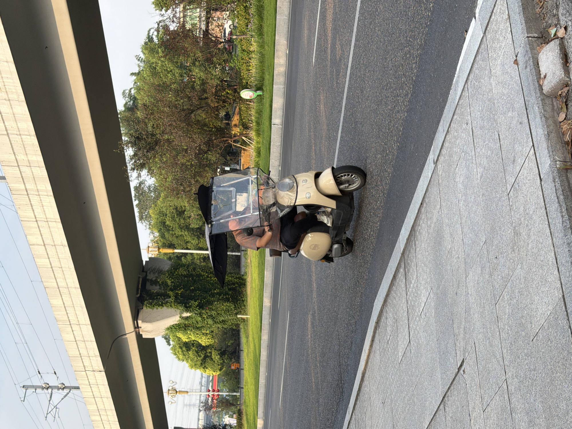 A man rides a three-wheeled electric scooter equipped with a transparent canopy and black umbrella for sun protection. The scooter is beige and has a single wheel in the front and two wheels in the back. The man is wearing a short-sleeved patterned shirt and dark pants. He is on a paved road with a grey sidewalk to the left. In the background, there is a large concrete overpass for what appears to be a railway or elevated road, with overhead power lines. Lush green trees and some vegetation line the roadside, and a yellow excavator is visible in the distance. The scene appears to be in an urban or suburban area in Jiangjin, China, possibly during daytime given the lighting.