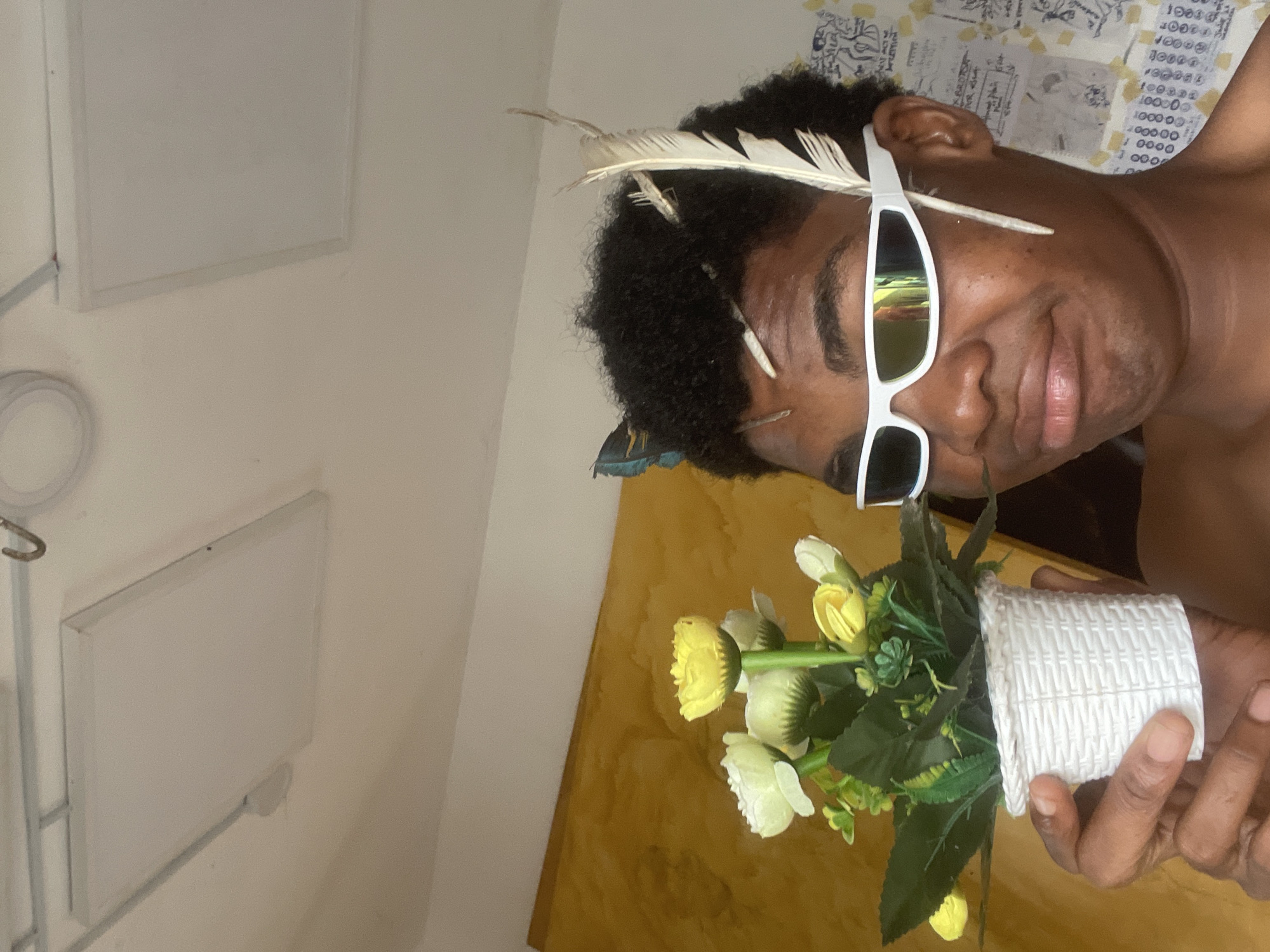 In Ado, Nigeria, a person with dark skin, wearing white-rimmed sports sunglasses, is pictured in an indoor setting. The individual is holding a small, white, woven pot containing a bouquet of artificial yellow and white flowers with green leaves. Several small objects, including what appear to be twigs and a feather, are placed in the person's dark, curly hair. The background features a textured yellow wall and a white ceiling with inset rectangular light panels. The overall scene is bright, with harsh lighting.