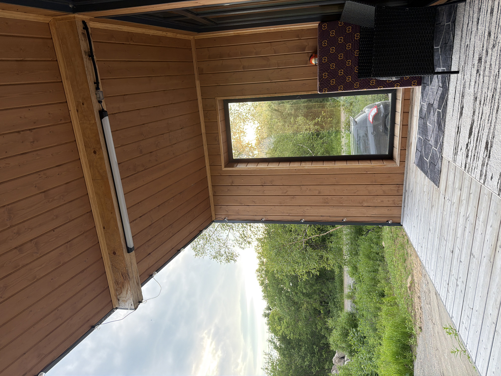 The image shows a porch of a wooden house with a view of a green forest. The porch is made of light-colored wooden planks. On the porch, there is a dark-colored wicker chair and a mattress with a repeating pattern. A window with a view of a forest and a dark-colored car is visible. The ceiling of the porch is made of wooden planks and has a light fixture. The sky is cloudy and the sun is setting. The location is the city of Novy, Russia.
