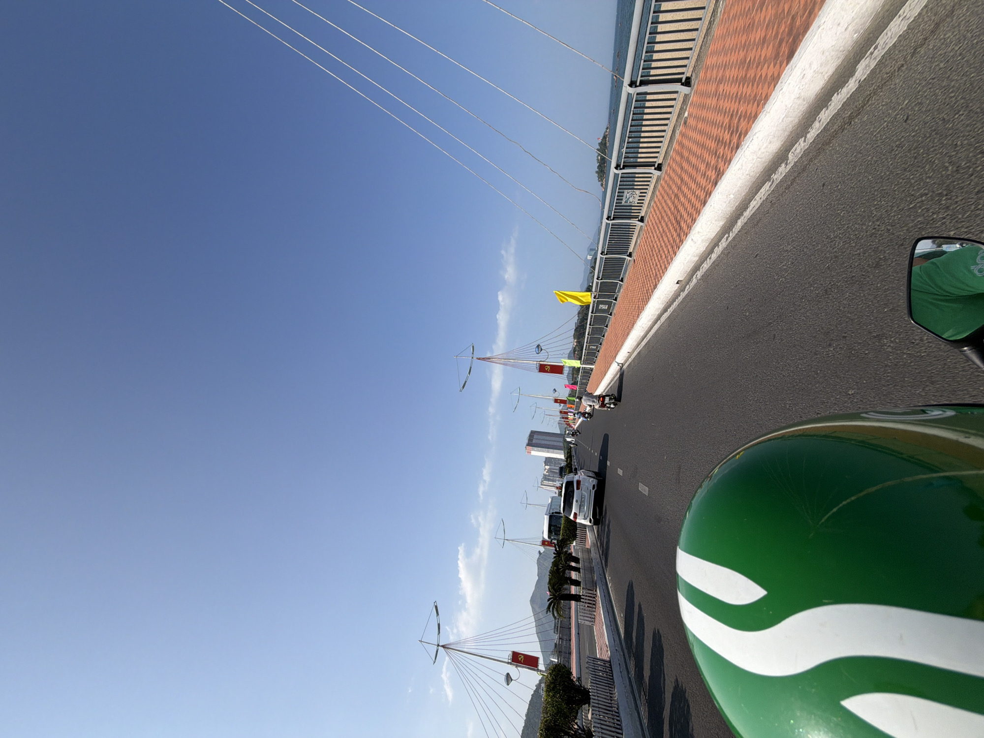 The image depicts a sunny day on a bridge in Nha Trang, Vietnam. The viewpoint is from the perspective of someone riding a motorcycle, with the green and white helmet prominently in the foreground. The bridge features a red tiled pedestrian walkway bordered by a metal railing and is lined with cable-stayed towers, some displaying red flags with yellow stars. Cars and other motorcycles are visible on the roadway. In the distance, there are buildings, mountains, and the ocean. The scene conveys a sense of urban transportation and coastal scenery.
