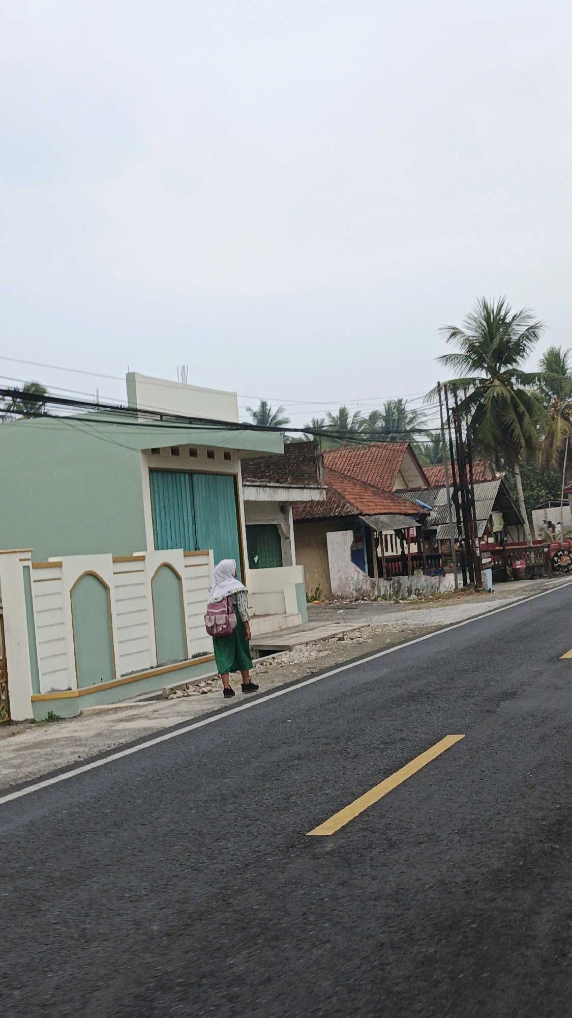 A young person wearing a green dress and a white hijab stands on the side of a paved road in Sukaresik, Indonesia. The person has a pink backpack on their back. The road has a yellow dashed line painted on it. To the left of the road is a light green building with decorative white trim. Behind the building and to the right are several other buildings with red tiled roofs and various structures. Palm trees are visible in the background, and the sky is overcast. The scene appears to be a residential or commercial area in a tropical climate.