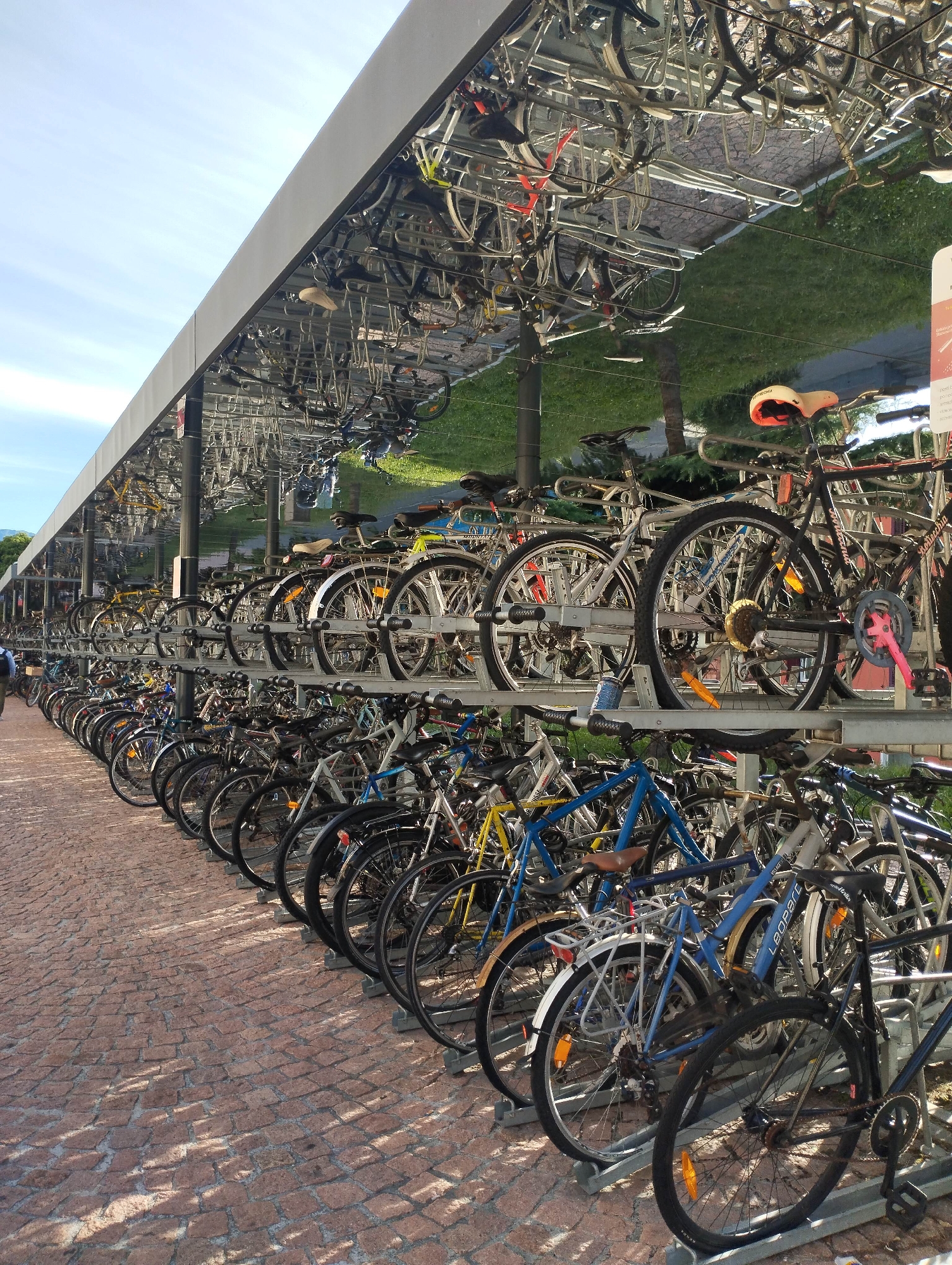 This image captures a large, multi-tiered bicycle parking facility under a bright, sunny sky. Rows of bicycles are neatly parked on both lower ground racks and elevated racks, creating a dense display of various bike types. The upper part of the parking structure features a reflective surface, possibly mirrored panels or highly polished metal, which vividly reflects the blue sky with wispy clouds, green grass, and trees from the opposite side, as well as the underside of parked bicycles.
The ground beneath the bikes is paved with reddish-brown cobblestones, casting dappled shadows from the bikes and the structure, suggesting it's either morning or late afternoon. Numerous bicycles of diverse styles are visible, including road bikes, mountain bikes, and city bikes, in a variety of colors such as blue, black, white, and yellow. Some bikes have accessories like fenders, baskets, and reflectors.
Notable text includes "LEOPARD" clearly visible on the frame of a blue bicycle and "GOSPEL" on another bike. A small, partially visible red and white sign stands on the far right, but its text is unreadable. On the far left, a person is partially visible, walking away from the bike racks. The overall scene depicts an organized, high-capacity solution for bicycle storage, likely in an urban or campus setting, emphasizing sustainable transportation. No specific activity beyond parking is occurring.
