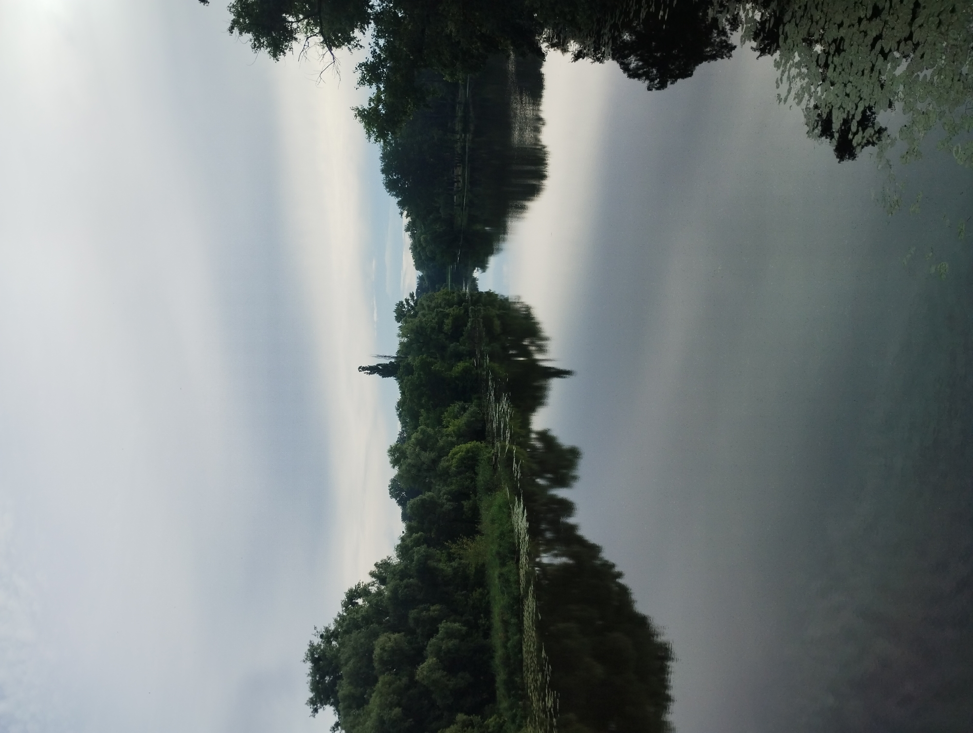 The image shows a tranquil river scene with lush green trees lining both banks. The water's surface is calm, reflecting the overcast sky and the dense foliage. In the distance, a prominent statue stands tall against the horizon, silhouetted by the sky. This statue is a historical monument located on the banks of the Psel River in the city of Sumy, Ukraine. The monument is a tribute to the defenders of the city, and it's a significant landmark, often referred to as the "Motherland" monument. The overall atmosphere is serene and natural, with the statue adding a touch of historical significance to the picturesque landscape.
