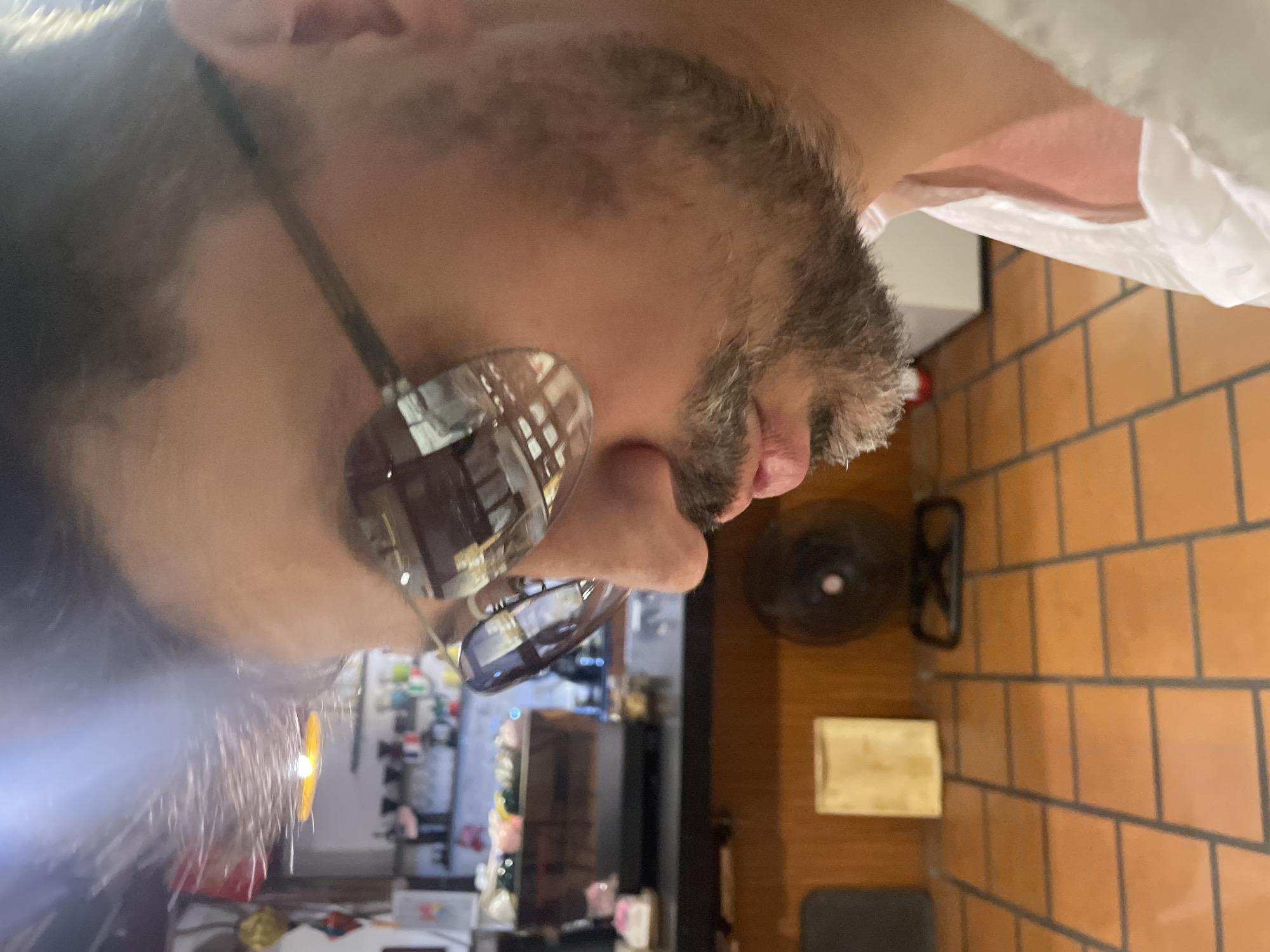 The image shows a close-up, upside-down shot of a person's face, with a focus on their chin, mouth, and stubble. They are wearing sunglasses with large, reflective lenses that mirror the surroundings, showing what appear to be shelves with bottles and possibly some decorative items. The person's skin is visible, along with areas of stubble and some darker hair on their upper lip and chin. A white, crinkled material is partially visible in the upper right portion of the frame, obscuring some of the background.
The background consists of a tiled wall with warm, orange-toned square tiles, suggesting an indoor setting. To the right of the person's face, a black fan with a circular grill is visible. Below the fan, a light-colored wooden block sits on the tiled surface. Further in the background, blurry shelves are visible, hinting at a retail or storage environment, possibly a shop or a home bar.
The lighting suggests it might be daytime, but the indoor setting makes it difficult to ascertain the exact time of day or weather conditions. The cropping and angle of the image create a somewhat abstract and intimate portrait, with the reflective sunglasses adding another layer of visual interest by capturing fragmented details of the environment. There is no visible text in the image.