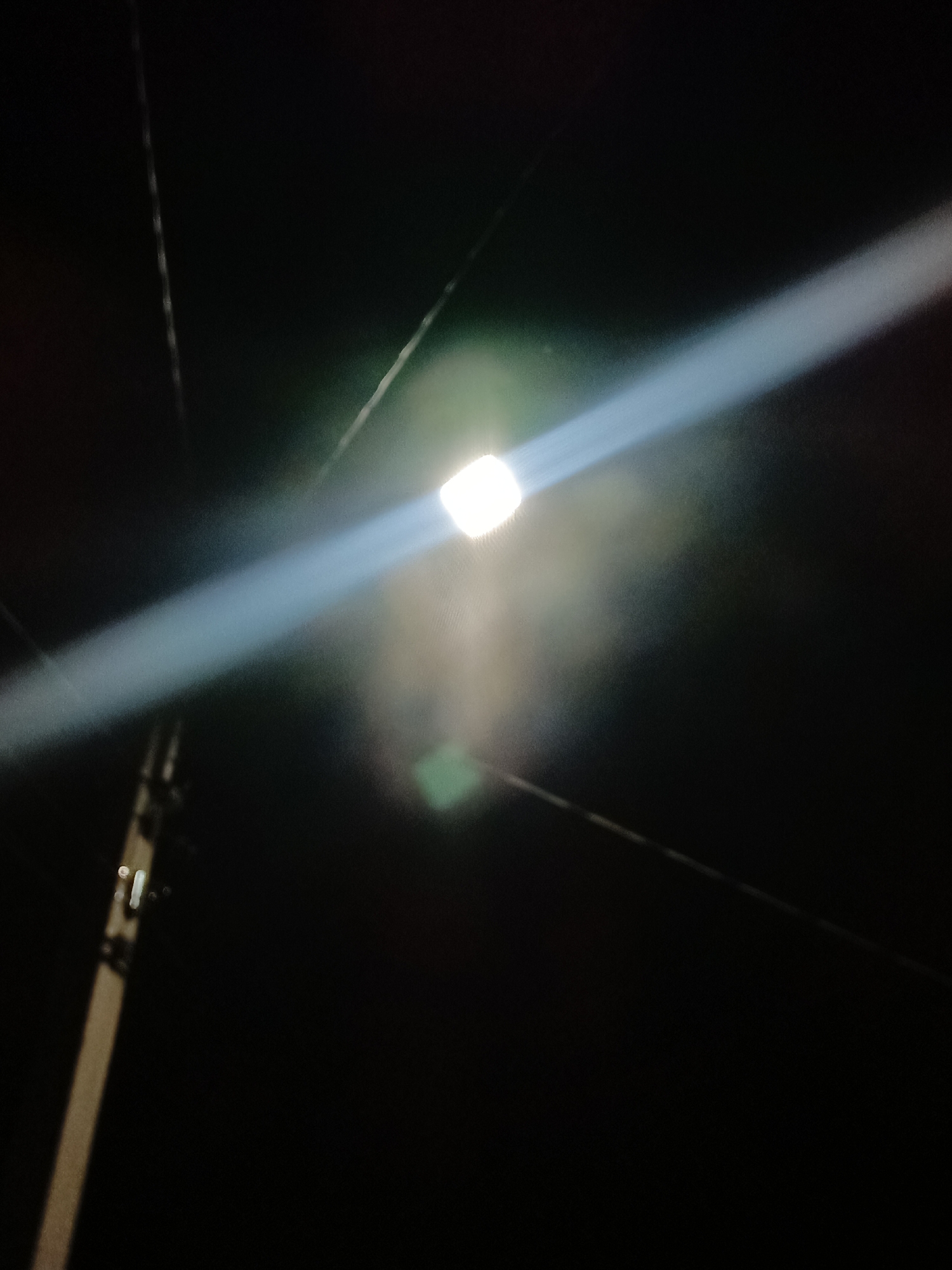 The image captures a streetlamp at night, casting a bright beam of light diagonally across the dark sky. The light source is a small, white, glowing orb. A portion of a utility pole, likely made of wood or concrete, is visible on the left side of the frame, with some attachments or insulators. The overall scene is dark, suggesting it is nighttime in an outdoor urban environment. There are no people or animals present in the image. The setting appears to be a typical street in Shymkent, Kazakhstan, characterized by street lighting and utility infrastructure.
