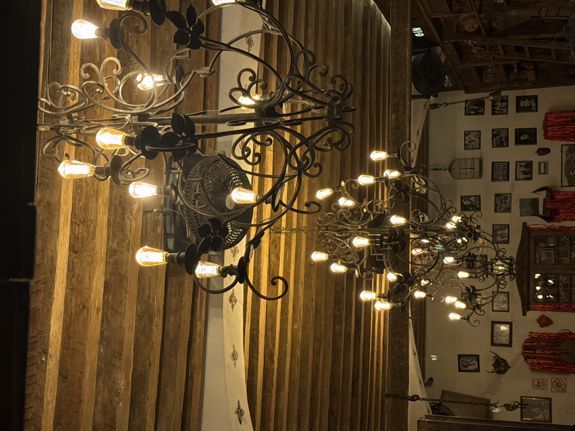 Two ornate, black metal chandeliers adorned with numerous glowing light bulbs hang prominently. The background consists of vertical wooden panels. A black electric fan is attached to the wall behind the left chandelier. The wall in the background has a collection of framed pictures and decorative items, including what appears to be a cupboard with glass doors displaying items. The scene is inside a restaurant in Puebla, Mexico.
