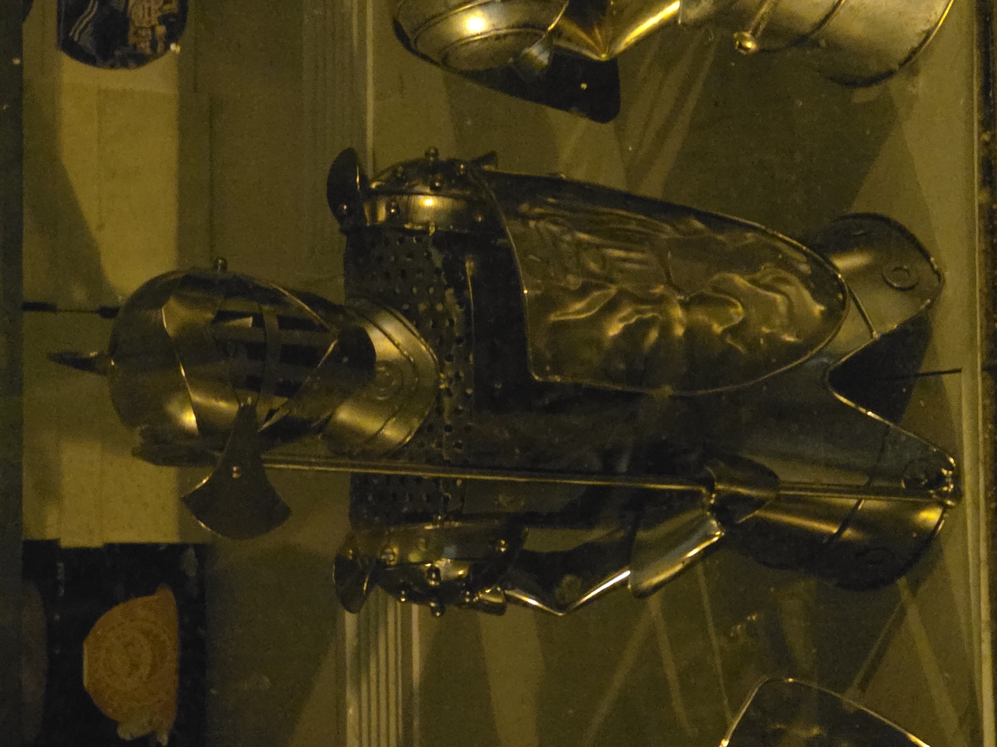 The image displays a collection of metallic armor pieces, likely historical or ornamental, within a dimly lit indoor setting. The central subject is a robust, dark metallic armor section, possibly a breastplate or torso guard. This piece features a heavily textured, almost studded surface in places, contrasting with smoother, highly reflective sections that show intricate curves and patterns, possibly depicting a stylized sun or emblem. Strong golden-yellow light illuminates these surfaces, creating bright highlights and deep shadows.
To the left, a more rounded, conical metallic object with visible slits suggests a helmet or another protective headpiece. Extending horizontally from this area is a slender metallic shaft with a small, flat, axe-like head at its end, possibly a weapon or decorative element. Other polished and curved metal forms, appearing to be additional armor components such as gauntlets or sections of leg armor, are visible scattered around the main display.
The background is a muted, light brownish-yellow wall or display case interior, with subtle textures. The lighting is artificial, casting a warm, golden hue over the entire scene, accentuating the metallic sheen of the armor. No people are visible, and the objects are presented as static exhibits, implying a museum, gallery, or private collection setting. There is no discernible text within the image. The orientation of the armor, lying horizontally, suggests it might be displayed flat or the image was captured at an oblique angle.