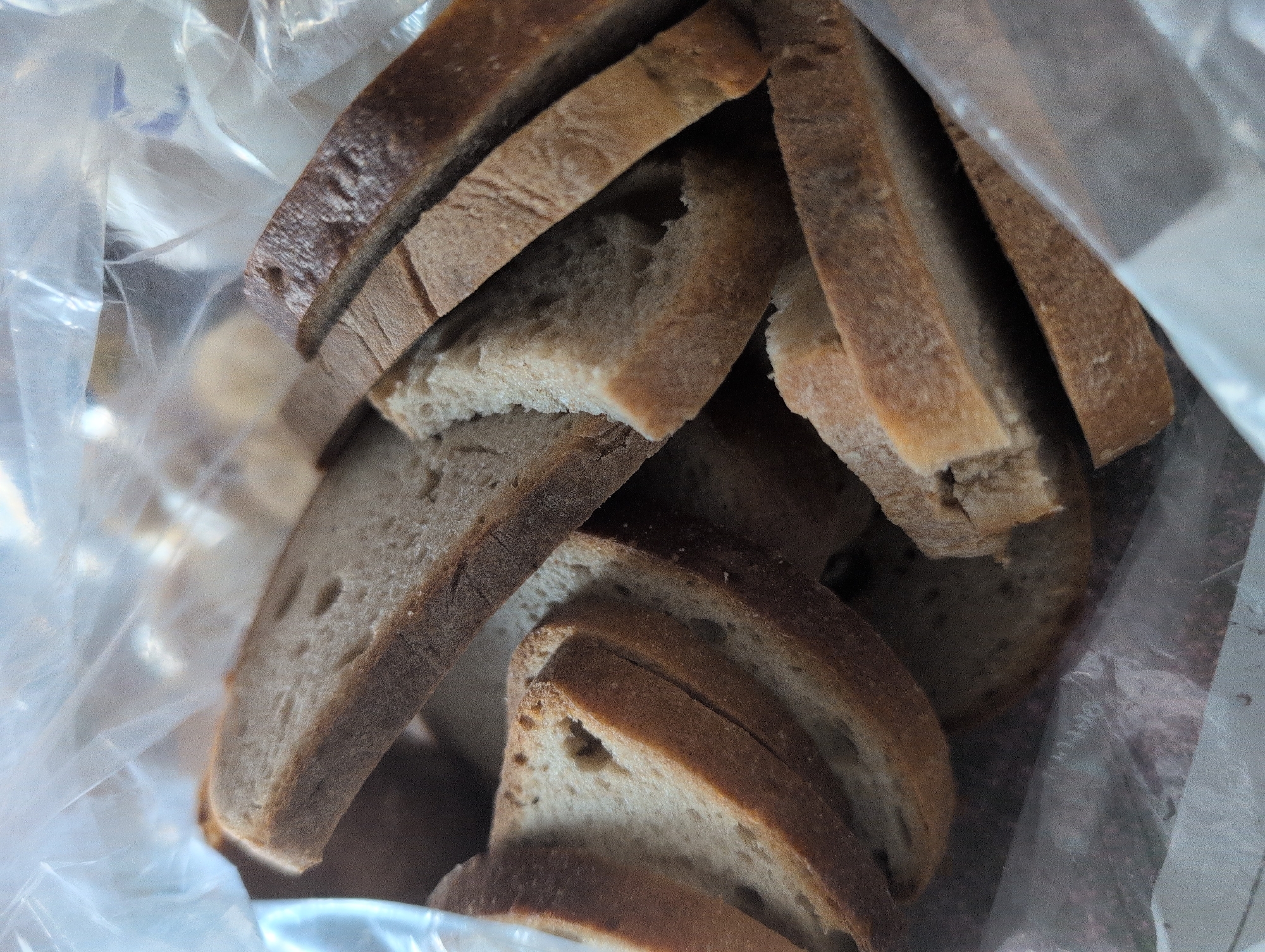 The image shows a close-up, overhead view of several slices of bread inside a clear plastic bag. The bread appears to be a darker, whole-grain variety, with visible crusts and a textured interior. The slices are stacked and angled within the bag, creating a layered effect. Some slices show signs of being slightly torn or unevenly cut. The plastic bag is slightly creased and reflects light in places, obscuring some of the details of the bread underneath. There are no people or discernible actions depicted in the image. The environment appears to be an indoor setting, but no specific location within Harsewinkel, Germany, can be identified from the visual information provided.