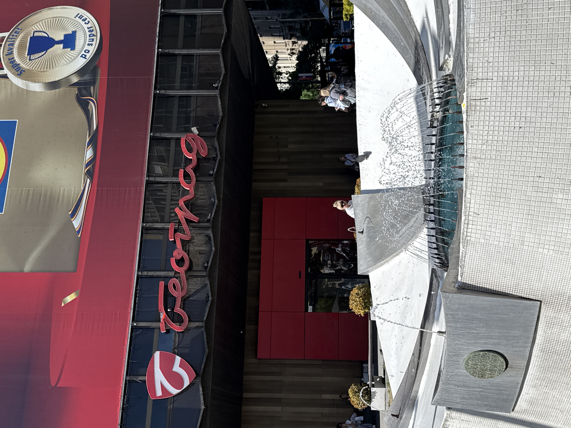This image captures a view of a city street in Vracar Urban Municipality, Serbia. The foreground features a modern fountain with water spraying from multiple jets into a semicircular basin. Pedestrians are visible walking along the pavement. Adjacent to the pavement is a building with prominent signage. The name "Tempo" is displayed vertically in red lettering, next to a white logo of a stylized number '3'. Above this, a large banner with the Lidl logo is visible. Further back, urban buildings and greenery can be seen along the street.
