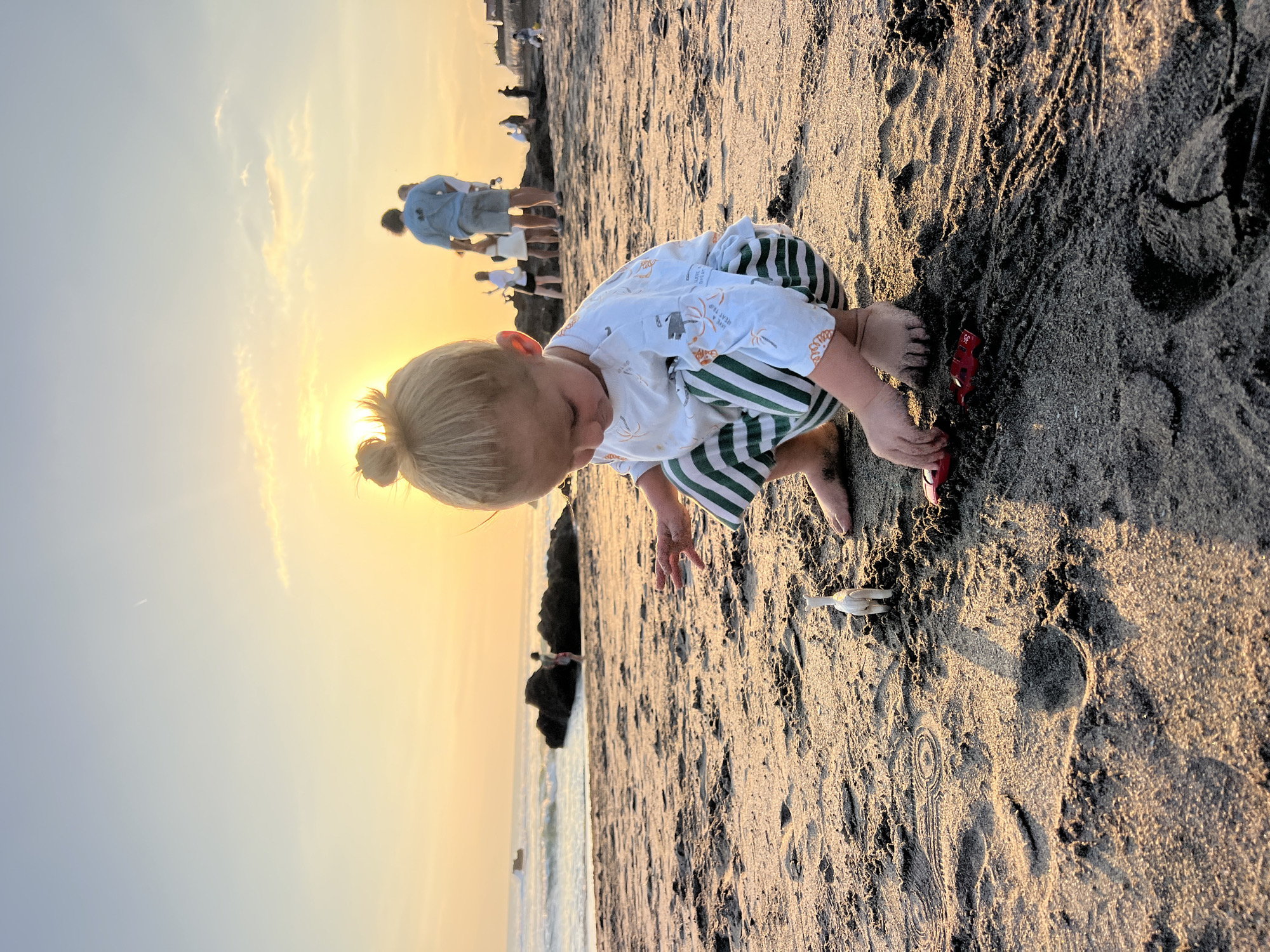 A young child with blonde hair in a bun is crouching on a sandy beach in North Kuta, Indonesia. The child is wearing a white t-shirt with orange palm tree prints and green and white striped pants. The child is playing with small toys, including a white animal figurine and a red toy car. The sun is setting, casting a warm golden glow over the scene. In the background, the ocean meets the shore with gentle waves, and several people are walking along the beach. The sand is textured with footprints and patterns, indicating a day of activity. The overall atmosphere is peaceful and serene, capturing a moment of childhood joy at sunset.