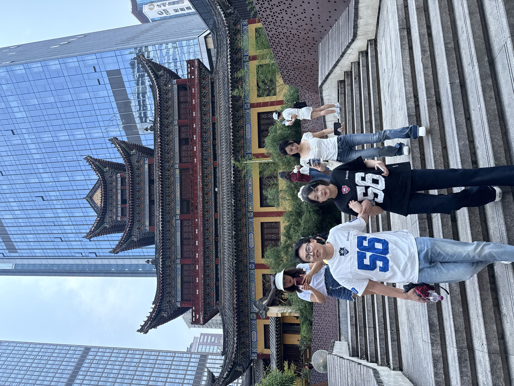 A group of young people is captured on a wide stone staircase in an urban setting, featuring a striking juxtaposition of traditional and modern architecture. The image is oriented vertically (appearing horizontally in the prompt, it should be rotated clockwise 90 degrees for correct viewing).
On the left, a grand, multi-tiered traditional Chinese building with elaborate eaves, intricate carvings, and vibrant red, blue, and gold accents stands prominently. Its dark tiled roofs and ornate wooden structures contrast sharply with the sleek, reflective glass facades of modern skyscrapers towering behind and beside it, one of which shows reflections of the sky and surroundings. A sign on a modern building in the background displays Chinese characters, including "星" (Star), "酒店公寓" (Hotel Apartment), "金" (Gold), and "会所" (Clubhouse).
The group consists of at least seven individuals, primarily young adults. In the foreground, a young man wears a white jersey with the number "95" in blue, along with "Battlefield" and an "HK" logo. He holds a clear plastic water bottle. Next to him, a young woman wears a black jersey with "86" in white, also featuring "Battlefield" and an "HK" logo. Other individuals in the group wear casual attire, including white t-shirts, jeans, and sneakers. Some are holding mobile phones, and one person wears a white visor.
The staircase itself is made of light grey, rough-hewn stone, leading upwards. Lush green foliage and smaller traditional architectural elements are visible between the main building and the staircase. The lighting is bright and even, indicating daytime with a clear sky, parts of which are visible between the buildings. The scene conveys a dynamic mix of historical culture and contemporary urban life, characteristic of places like Yuzhong District, China.