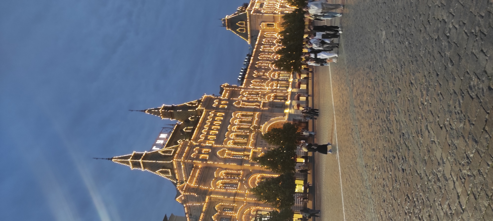 A low-angle, vertical shot depicts the GUM department store on Red Square in Moscow, Russia, illuminated by numerous string lights against a twilight sky. The architecture is ornate, with multiple towers and arched windows. A group of people, appearing to be tourists, are walking across the cobblestone square, some in the foreground and others further away near the building. The lighting creates a festive atmosphere.