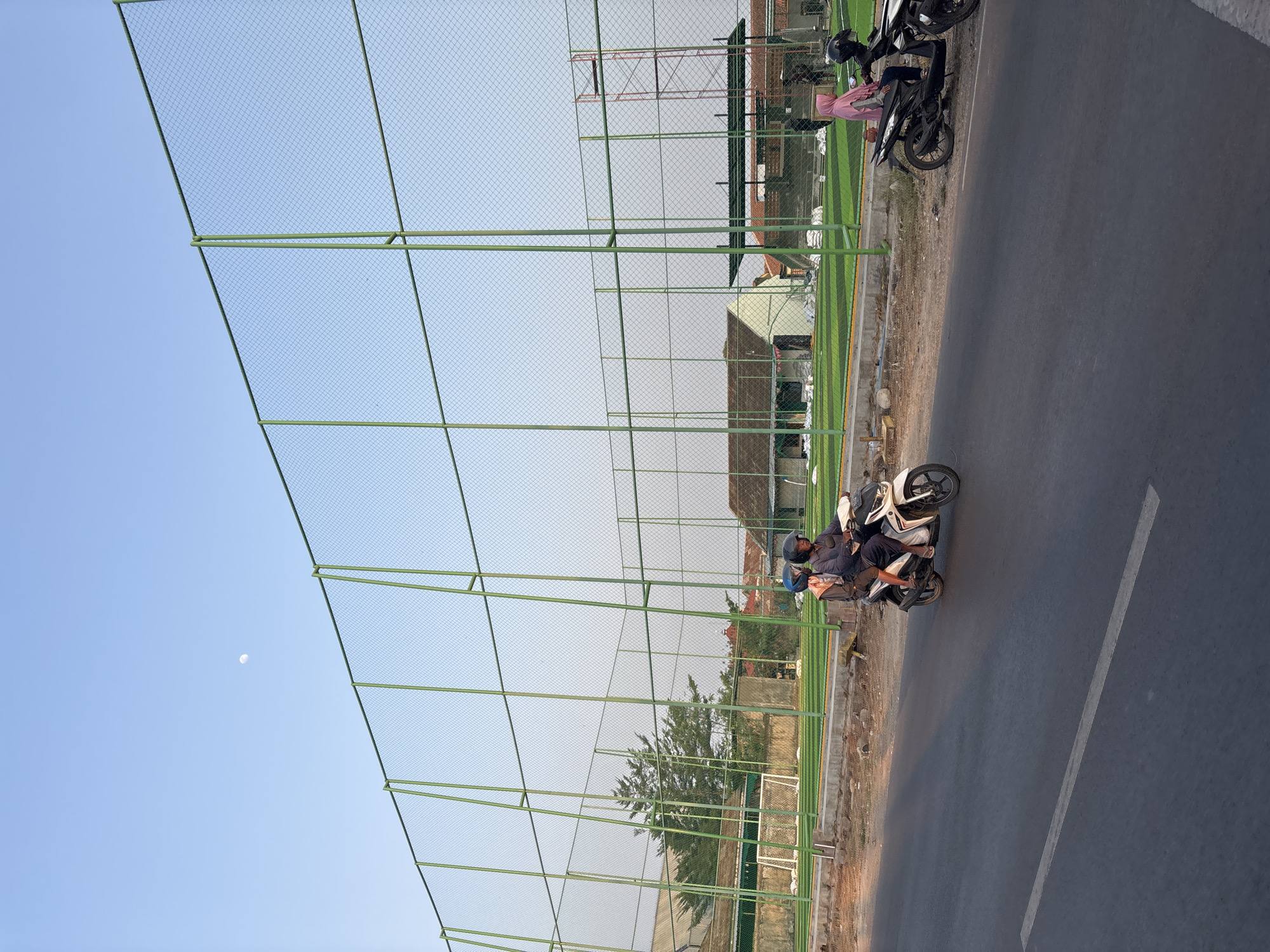 The image captures a street scene in Cirebon, Indonesia, likely near a sports facility. In the foreground, a paved road with a white lane marker runs horizontally. Two motorbikes are visible on the road, one white and one black. The white motorbike has two people on it, a driver wearing a helmet and a passenger behind. The black motorbike is parked on the side of the road, with a person wearing a pink hijab seated on it.
Behind the road, a green fence encloses a large area with a bright green artificial turf field, characteristic of a soccer or futsal field. The field has white lines marking boundaries and a goalpost visible on the left side. Several small white bags are scattered around the field's edge, possibly for construction or maintenance materials. Buildings with tiled roofs are visible beyond the field, including a light-colored structure with a dark green roof overhang. A tall red scaffolding structure stands to the right of the field.
The sky above is a clear, pale blue, with the moon visible as a small, pale disc. The overall lighting suggests it is late afternoon or early evening. The environment appears to be a mix of urban infrastructure and sports amenities.