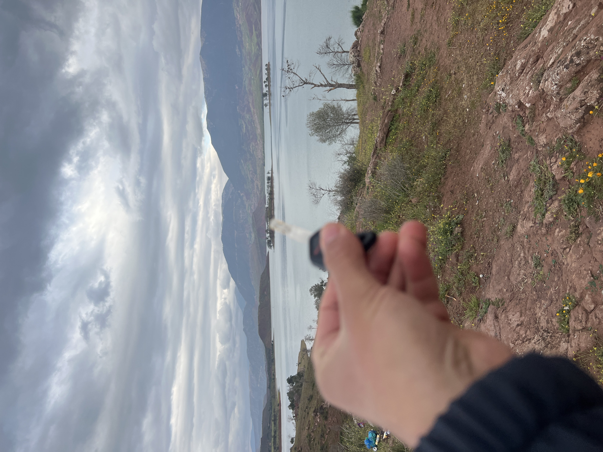 A landscape view, rotated clockwise, shows a body of water surrounded by mountains and land. The sky is cloudy. In the foreground, a person's hand is holding a car key. The ground is rocky with some vegetation. The location is Bin El Ouidane, Morocco.