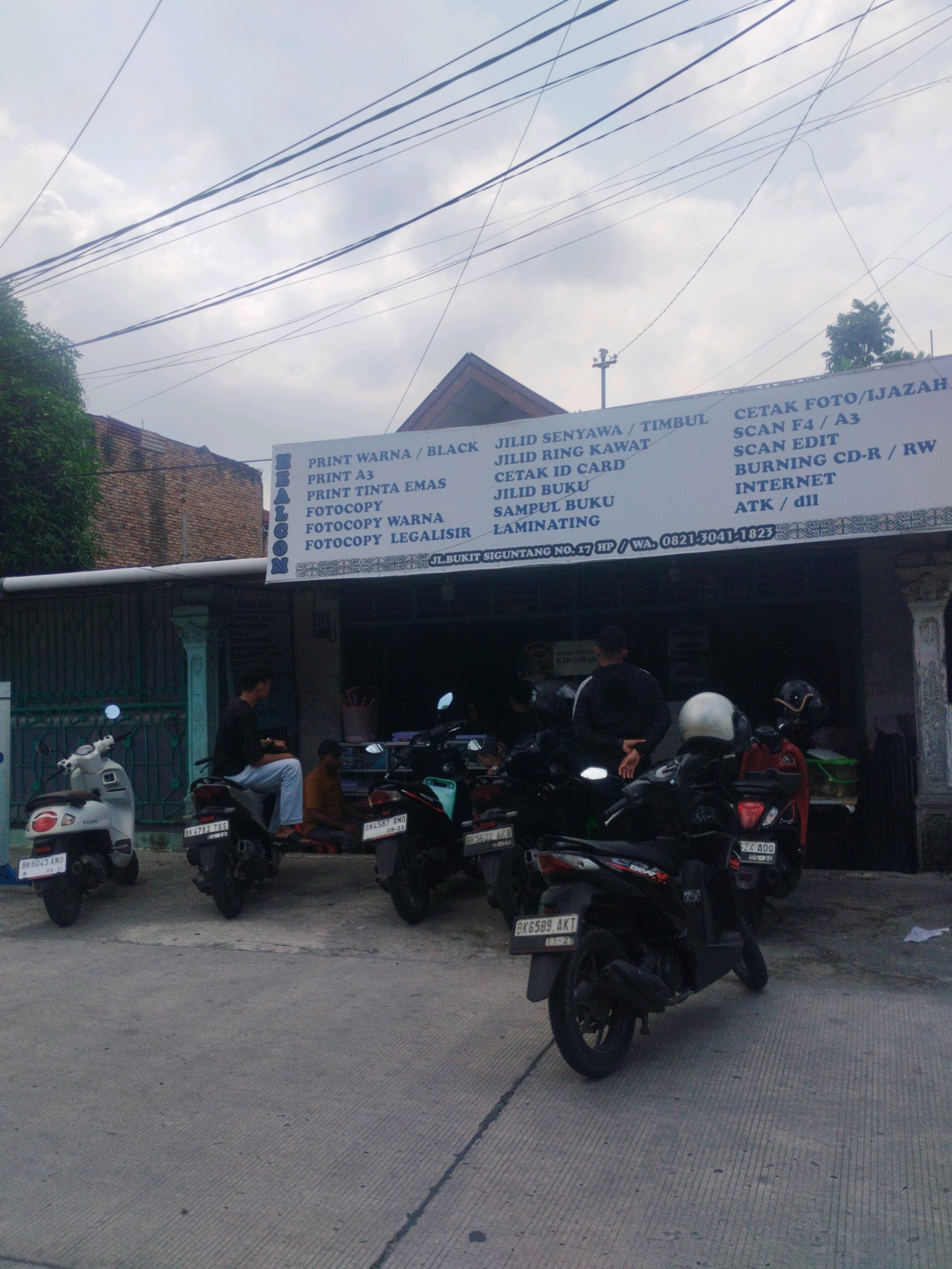 The image shows the exterior of a shop in the City of Medan, Indonesia, as indicated by the license plates on the motorcycles. The shop appears to be a print and copy service. A banner above the shop entrance lists various services offered, including "PRINT WARNA / BLACK", "JILID SENYAWAL / TIMBUL", "CETAK FOTO/IJAZAH", "PRINT A3", "JILID RING KAWAT", "SCAN F4 / A3", "PRINT TINTA EMAS", "CETAK ID CARD", "SCAN EDIT", "FOTOCOPY", "JILID BUKU", "BURNING CD-R / RW", "FOTOCOPY WARNA", "SAMPUL BUKU", "INTERNET", and "FOTOCOPY LEGALISIR". The banner also displays the address: "JL. BUKIT SIGUNTANG NO. 47 HP / WA. 0821-3041-1823".
Several motorcycles are parked in front of the shop. Two individuals are visible outside the shop. One person is seated on a motorcycle, wearing a black shirt and blue pants. Another person is seated on the ground near the entrance. The sky is overcast. The environment suggests a commercial area with modest infrastructure. The overall scene captures a typical day at a local business in Medan.