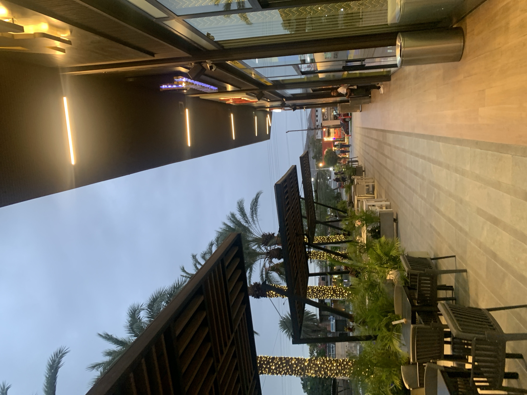 This image captures an outdoor commercial promenade in Pampatar, Venezuela, during dusk or early evening, as indicated by the overcast sky and the activation of various artificial lights.
On the left, a modern building facade features expansive glass windows reflecting the sky, below which a dark wall is illuminated by several warm, vertical linear light fixtures. A blue and purple neon sign is visible through the glass storefront.
The right side of the frame showcases a wide, light-toned paved walkway, marked by a darker central strip, extending into the distance. This area is furnished with numerous dark-colored outdoor tables and chairs, arranged for dining or lounging. Green plants in planters and several tall palm trees are interspersed among the seating arrangements. Many of the palm tree trunks are decorated with warm-toned string lights, contributing to the evening ambiance.
In the mid-ground, a tall, cylindrical stainless steel trash receptacle stands near the building. Several people are visible; one individual in a white t-shirt and light pants stands by the building, and others are seen further down the promenade, suggesting a leisurely atmosphere.
The background reveals a street scene with additional buildings and vehicles. Faintly discernible text on distant building signs includes "Casani" and "PRIMA CAFÉ." The overall scene suggests a vibrant, well-maintained commercial and leisure space.