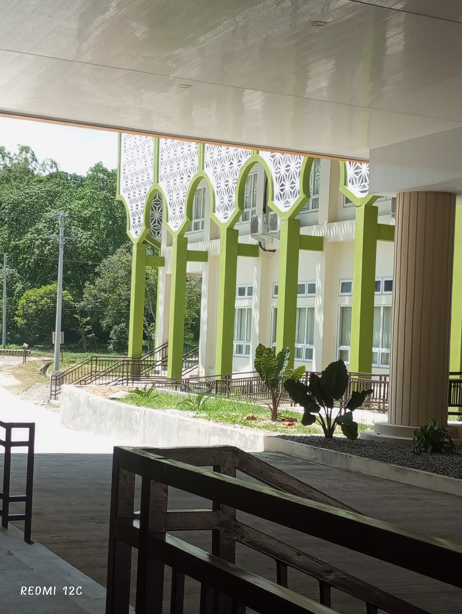 The image shows a view under a sheltered walkway facing a two-story building in Lhokseumawe, Indonesia. The building has white walls with green trim and is adorned with decorative arches and windows. A well-maintained garden with various plants is located in front of the building. Railings are present on the walkway and around the garden area. Beyond the building, there is a view of trees and a utility pole. The location is likely a public or institutional setting, possibly a school or government building in Lhokseumawe. There are no people visible in the scene.