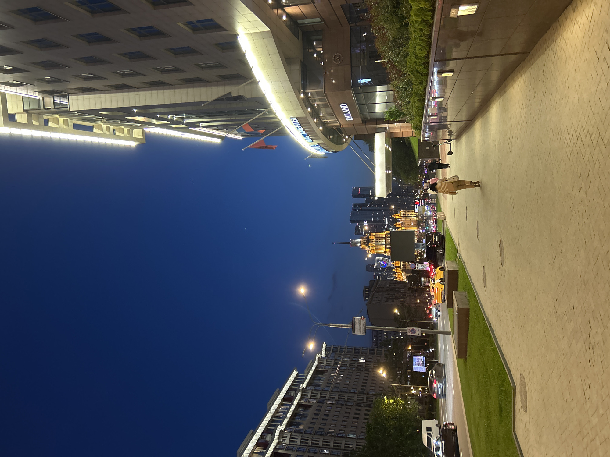 The image depicts an urban nighttime scene. A tall building with illuminated signage is visible in the foreground, with the inscription "SWISSÔTEL KRASNYE HOLMY MOSCOW". In the background, a cityscape with various illuminated buildings and structures is visible. A paved walkway extends into the scene, and people are walking along it. The sky is a deep blue. Streetlights and traffic are present.