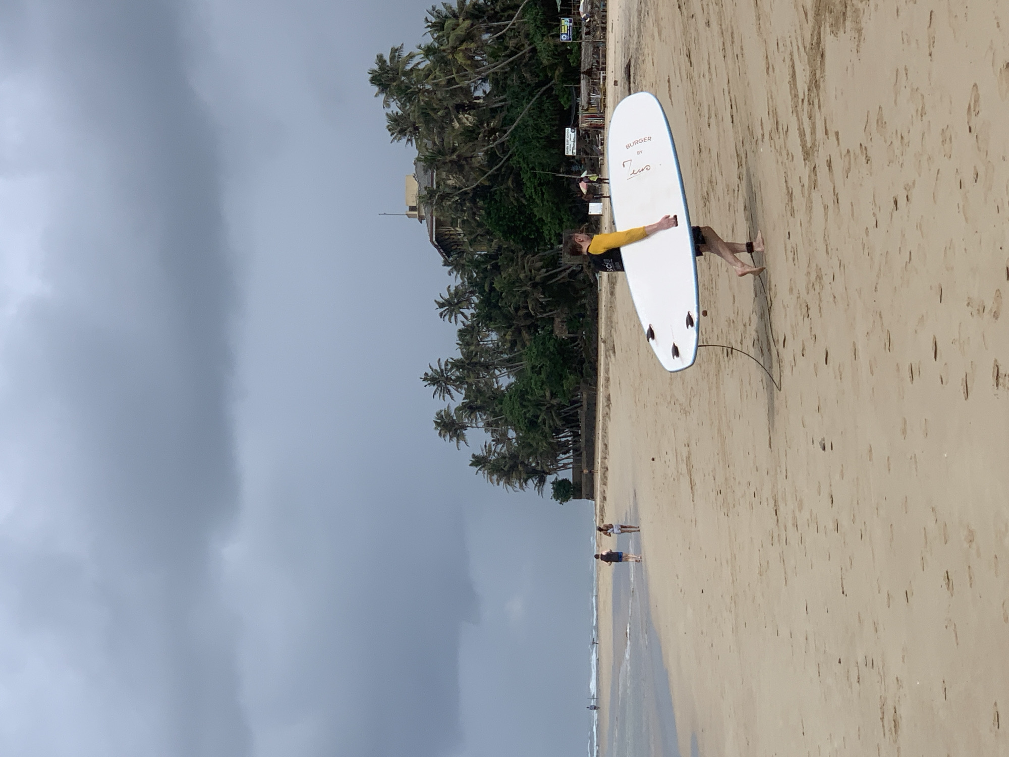 A surfer carries a white surfboard on a beach in Kathaluwa, Sri Lanka, under an overcast sky. The board, labeled "Burger by Zeus," is held horizontally by the surfer who is wearing a yellow long-sleeved shirt and dark shorts. The beach is tan with visible foot prints. In the distance, two people walk along the shoreline. Palm trees are visible in the background. The ocean is in the middle ground with people in the water.