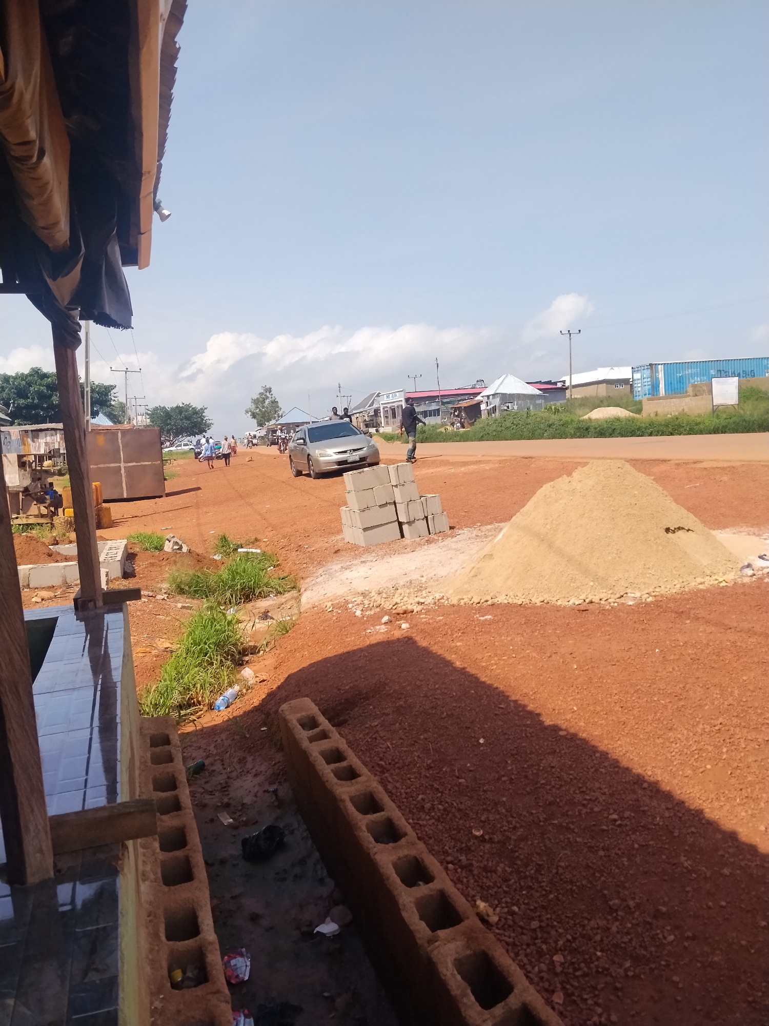 The image depicts a roadside scene in Durumi, Nigeria, characterized by a red dirt road and construction materials. In the foreground, to the left, a partially constructed building with tiled flooring and wooden supports is visible. Adjacent to it are two rows of concrete blocks, one filled with dirt and the other empty. A pile of sand is situated to the right of the road, with a stack of grey concrete blocks next to it.
The road itself is trafficked by a silver sedan. Several individuals are walking along the road in the middle ground. In the background, structures of various types are present, including buildings with corrugated metal roofs and shipping containers. Overhead, utility poles with wires are visible against a sky with scattered clouds. The overall environment suggests an active area with ongoing development or commercial activity.