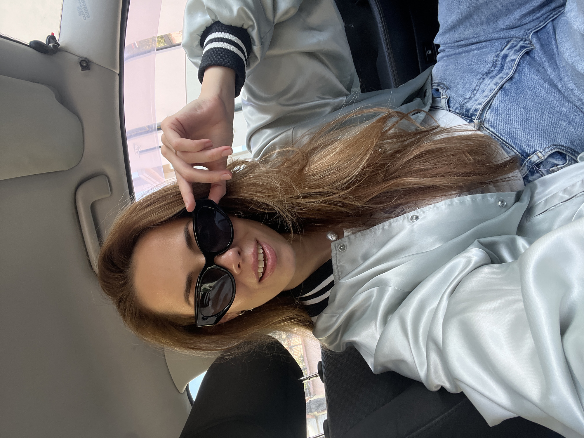 A female subject is observed in the front passenger seat of a vehicle, capturing a self-portrait. She has long, light brown hair and is wearing black cat-eye sunglasses, a light green satin bomber jacket with black and white striped cuffs, a white undershirt, and blue denim jeans. A ring is present on her right ring finger, and her right hand is positioned near the right temple of her sunglasses. She exhibits a slight open-mouthed smile while looking directly at the camera. The vehicle's interior includes a gray fabric passenger seat, a dark headrest, a gray headliner, and an A-pillar featuring 'CURTAIN AIRBAG' text. The exterior environment, visible through the tinted passenger window, shows indistinct buildings and foliage. This scene is located in Šušanj, Montenegro.