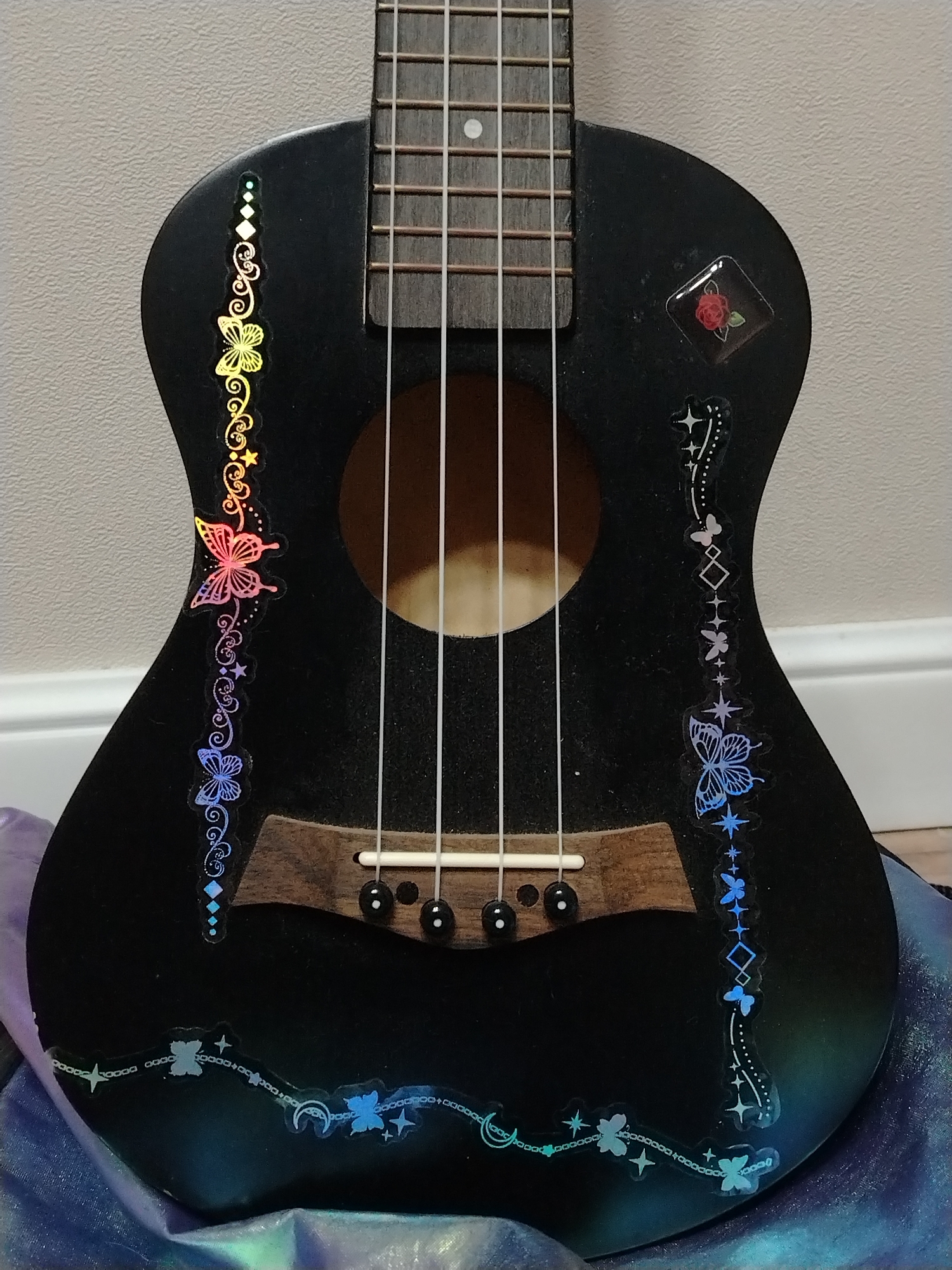 A close-up shot features a black ukulele adorned with colorful stickers. The ukulele is positioned vertically in the frame. Rainbow-colored butterfly and floral stickers run along the left side of the ukulele's body. On the right side, a series of blue butterfly and star stickers cascade down the body. A red rose sticker in a square frame is visible near the top right. A chain sticker with moons, stars, and butterflies runs along the bottom of the ukulele. The instrument has four strings, and the sound hole is visible. The ukulele rests on a shimmering purple and blue fabric. The background is a plain, light-colored wall and white baseboard. There is no indication in the image that it is located in Izhevsk, Russia.