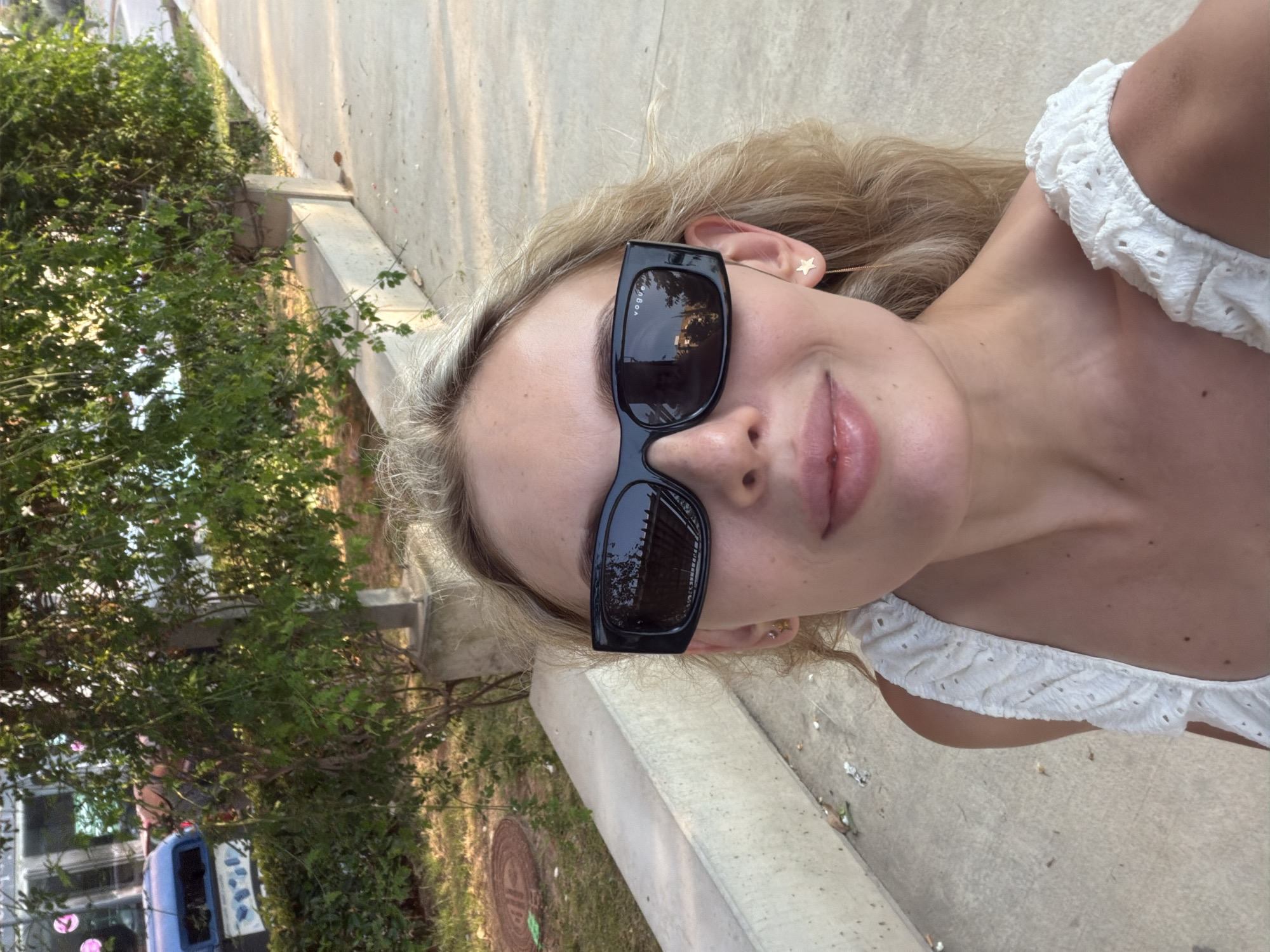 A young woman with blonde hair and fair skin is taking a selfie outdoors. She is wearing black sunglasses and a white, ruffled top. Her lips are slightly parted, and she appears to be smiling. A star-shaped earring is visible on her right ear. The background shows a concrete path next to some greenery and trees. In the distance, a light blue vehicle with some signage is visible, partially obscured by the foliage. The lighting suggests it is daytime. The image is oriented vertically, with the woman's face taking up a significant portion of the frame. There is no visible text in the image.