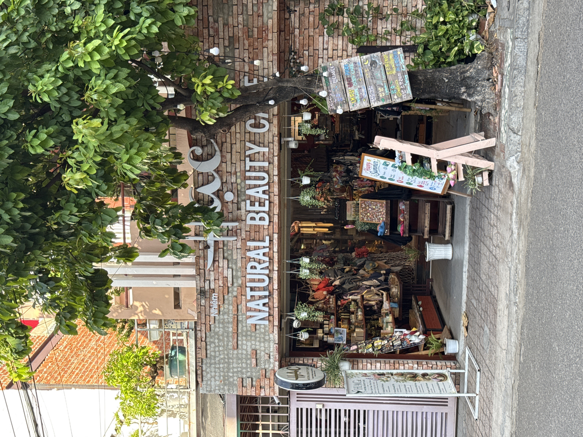 The image shows the storefront of a business named "HAC Natural Beauty Care" located in Nha Trang, Vietnam. The building features a brick facade with a sign that reads "HAC" and "NATURAL BEAUTY CARE" in prominent white lettering. The shop appears to sell various beauty products, bags, and accessories, displayed on shelves and hanging from the ceiling. A large tree with green foliage is situated in front of the store, partially obscuring the view of the entrance. String lights are visible hanging from the tree branches. Several signs, some with Korean text, are displayed outside the store, including one advertising "Happy Summer". The storefront is on a paved street, with a sidewalk in front. The overall scene suggests a commercial establishment in a tropical urban environment.