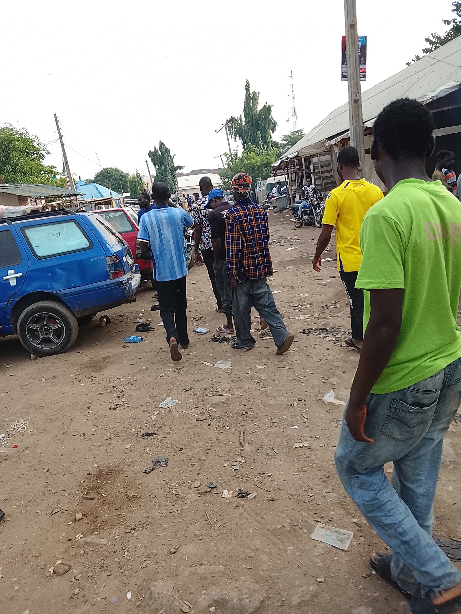 The image depicts a street scene in Abuja, Nigeria. Several people are walking along a dirt road littered with trash.
* **People:** About seven individuals are visible. One person is wearing a bright green shirt and faded blue jeans, walking towards the right. Another is wearing a yellow shirt. Others wear blue, plaid, and patterned shirts.
* **Objects:** A blue station wagon is parked to the left. Other vehicles, including motorcycles, are visible in the background. A utility pole with a banner is on the right.
* **Environment:** The road is unpaved and dirty. Buildings and trees are visible in the background.
* **Actions:** People are walking down the road.
* **Interactions:** The people appear to be walking together.