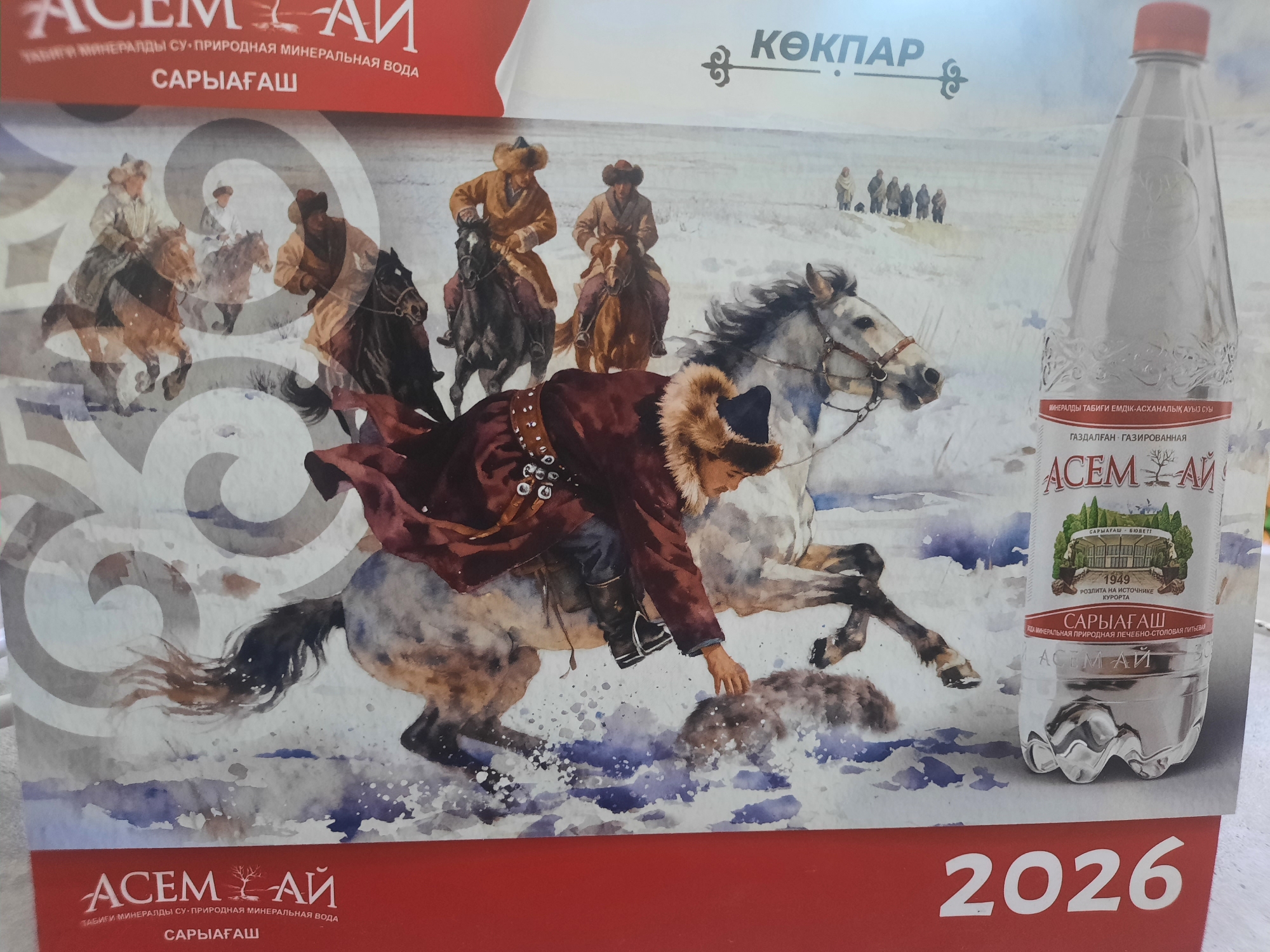 The image is a promotional calendar page for "ASEM-AI" mineral water. The calendar features a watercolor illustration depicting a group of individuals on horseback riding through a snowy landscape, possibly in a historical or traditional setting. In the foreground, a person in a fur-lined coat and hat is riding a grey horse, reaching down towards the snow. Behind them, other figures on horseback are visible in the distance. A bottle of "ASEM-AI" mineral water with a label that reads "SARIAĞAŞ" is prominently placed on the right side of the calendar page. The background depicts a snowy, mountainous or hilly terrain with a group of people standing in the distance. The bottom of the calendar page prominently displays the year "2026". The text on the calendar page is in Russian and Kazakh, referring to "ASEM-AI" and "SARIAĞAŞ", which is a resort town in the East Kazakhstan Region, not Zhanaozen.